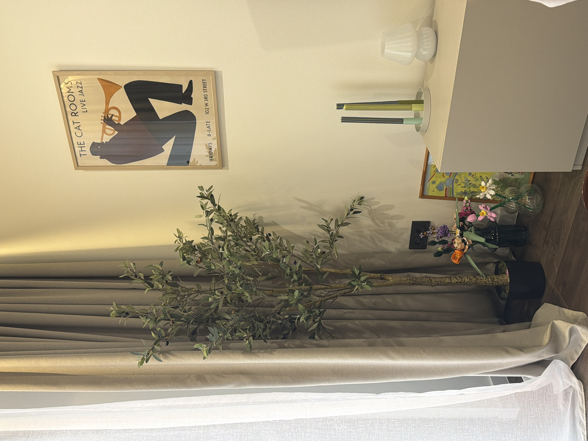 A still-life photograph presents an interior scene. On the left, a framed poster bearing the words "THE CAT ROOMS LIVE JAZZ" and an illustration of a figure playing a horn is mounted on a light-colored wall. Below, an olive tree in a black pot extends horizontally across the lower half of the image, its branches overlapping with heavy, light-tan curtains. At the bottom, white sheer curtains filter light, creating a softer, lighter area. To the right, a tall, light-gray cabinet stands adjacent to a collection of floral arrangements in vases. The color palette is dominated by neutrals and muted tones, with the only pops of color coming from the flowers and the poster.