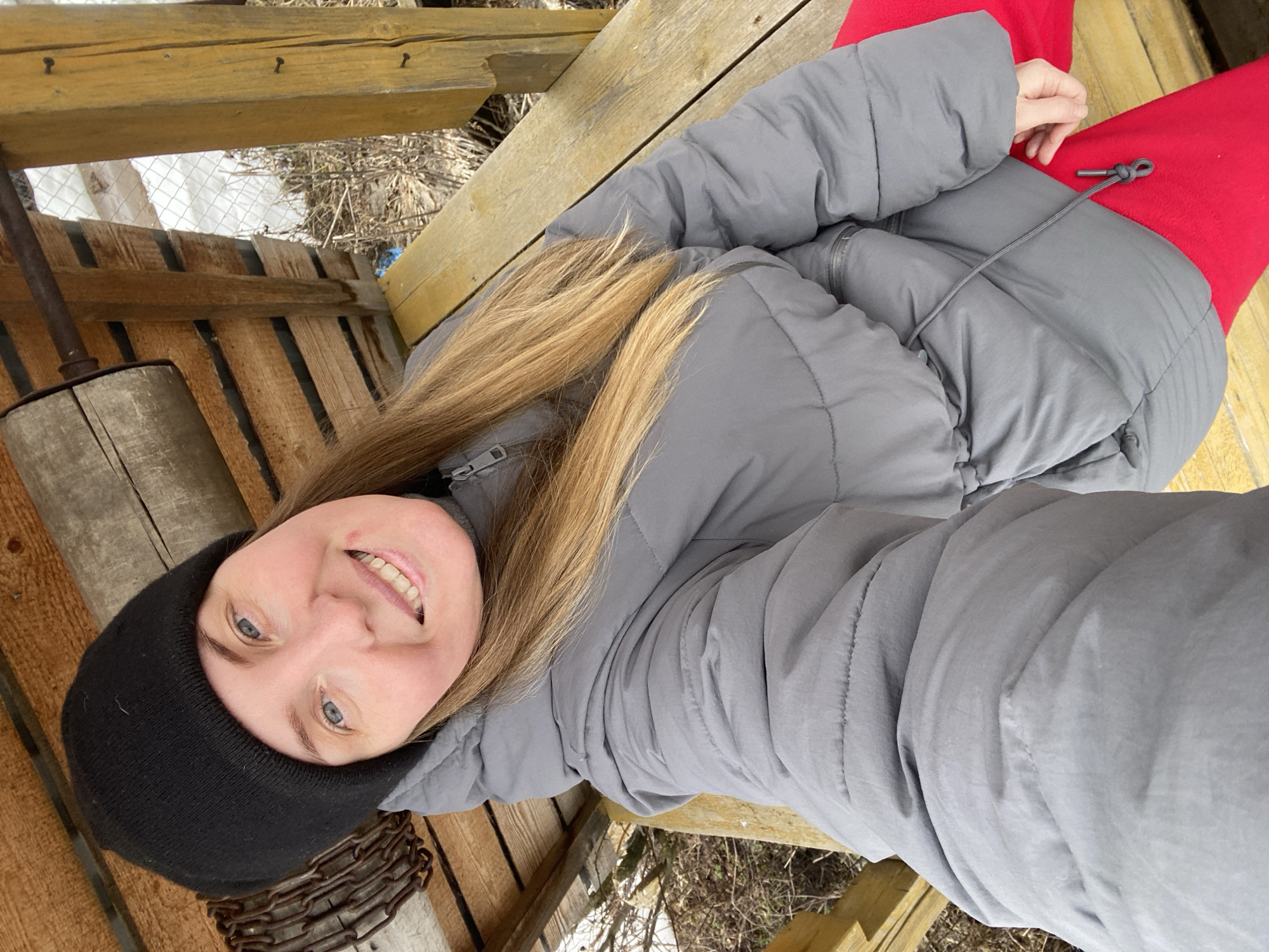 A woman with blonde hair wearing a black beanie and a gray puffer jacket over red pants smiles at the camera. She is positioned within a wooden structure, possibly a well or shelter, with exposed wooden planks. A metallic chain and a wooden cylinder are visible near her face. Outside the structure, snow-covered ground and bare vegetation can be seen. The image was taken outdoors in Chaikovsky, Russia, since this was a query criteria.