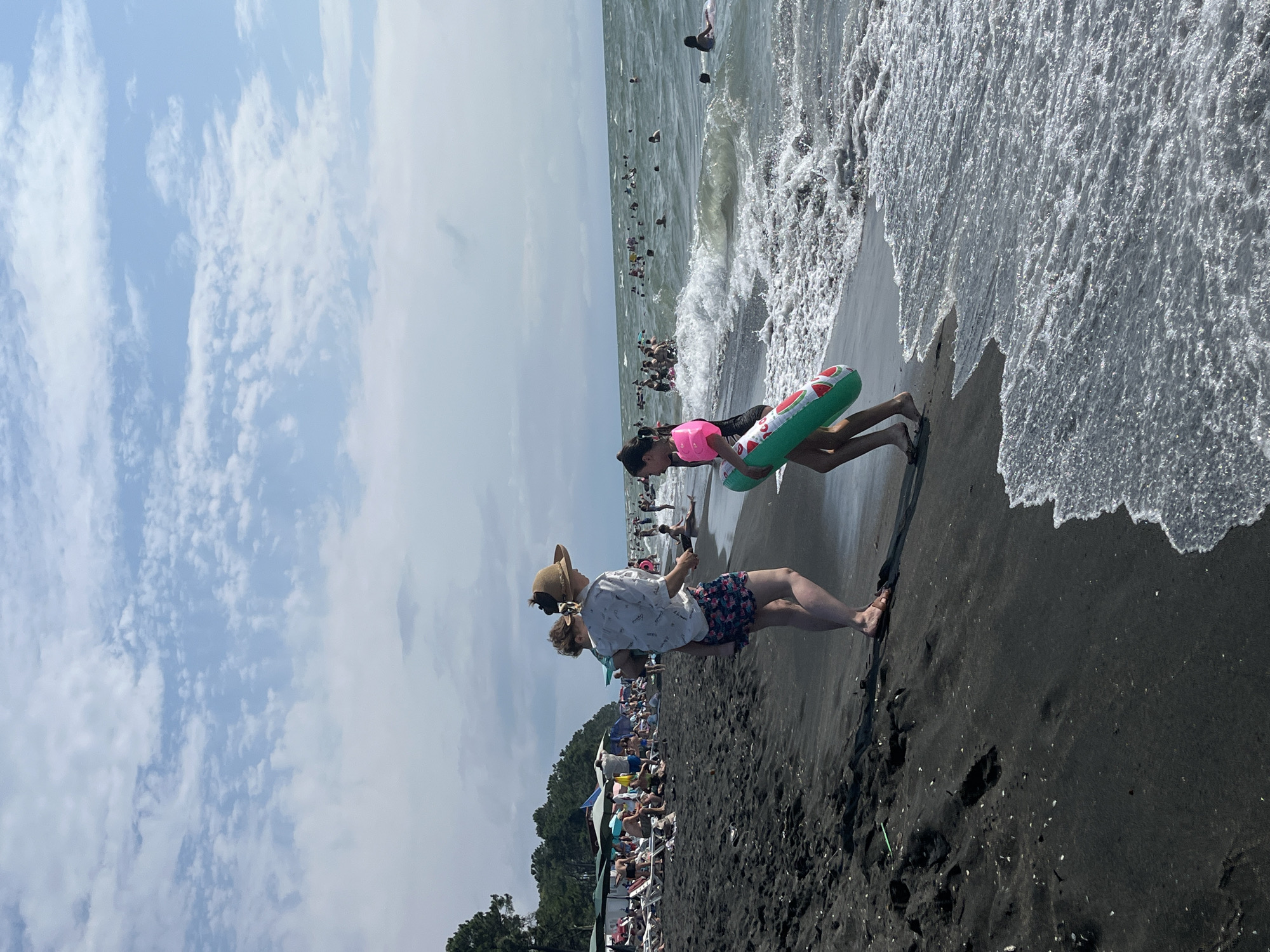 In Ureki, Georgia, on a sunny day, a beach scene unfolds with a dark sand shoreline meeting a wavy sea. The sky is partly cloudy with cumulus formations. In the foreground, a person wearing a straw hat, a white short-sleeved shirt with a pattern, and patterned shorts stands holding a child. Beside them, a young girl walks towards the sea, carrying a green inflatable ring with red watermelon slices. In the background, numerous people are visible on the beach, with some lounging on chairs under umbrellas and others in the water, swimming and enjoying the sea. Trees line the back of the beach.
