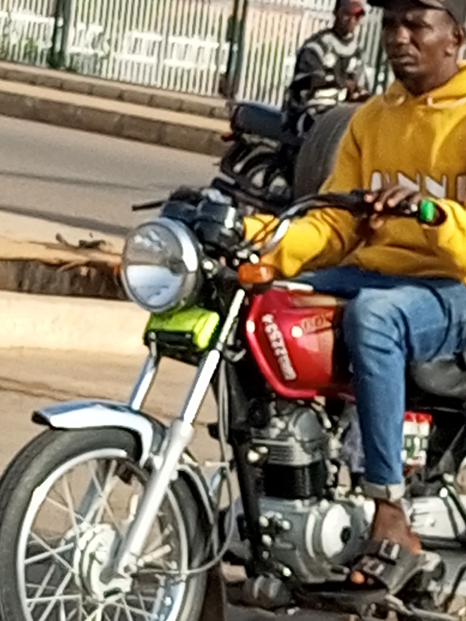 A man in a yellow hooded sweatshirt and blue jeans is riding a red motorcycle. He is wearing a dark baseball cap. The motorcycle has a silver front fender and handlebars, and a round headlight. There is another person on a motorcycle behind him, wearing a striped sweater and a dark cap. They are on a paved road with a sidewalk and a metal fence in the background. The scene appears to be in Tumfure, Nigeria.
