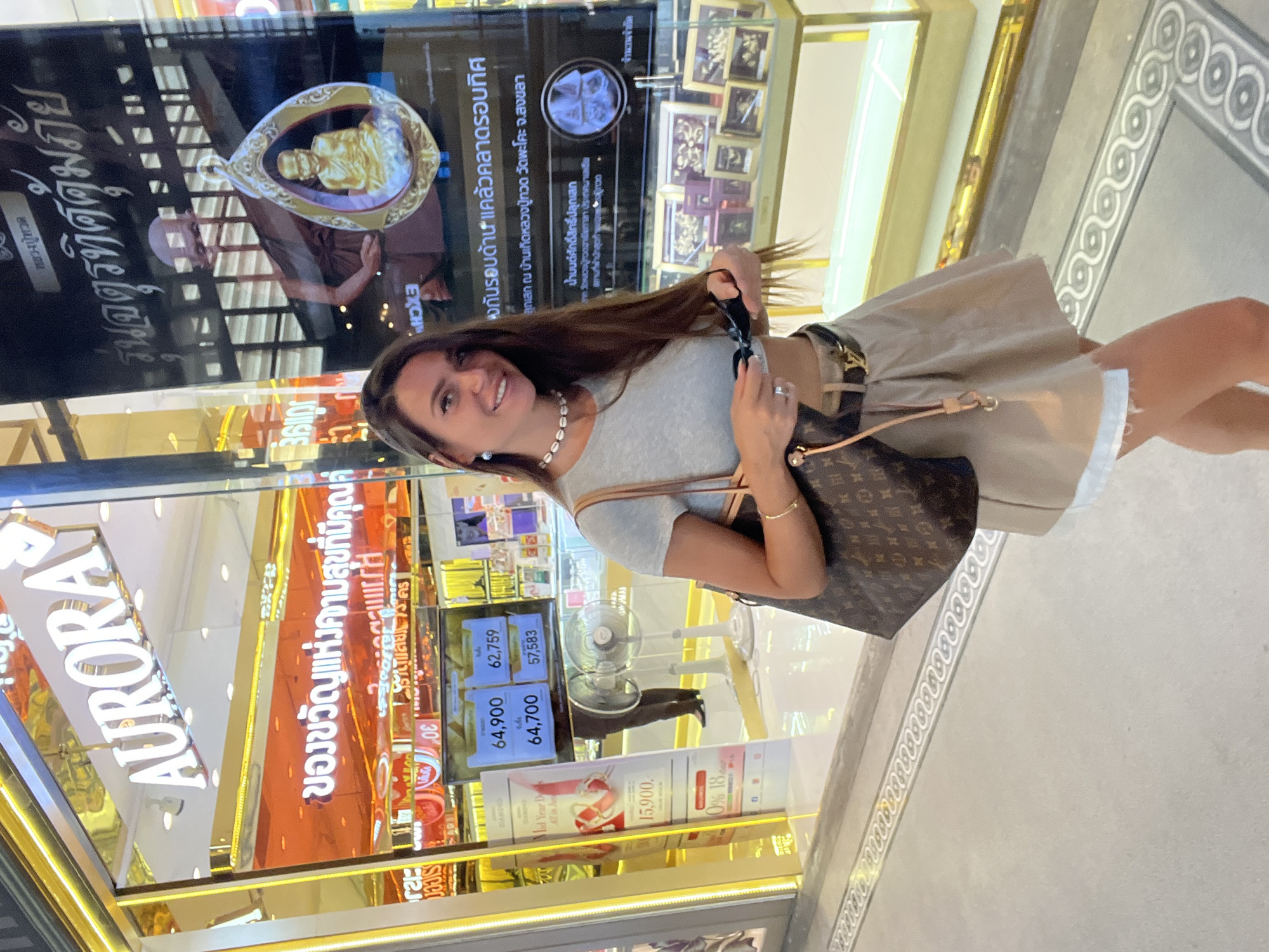 A woman with long brown hair stands in front of a store with a prominent "AURORA" sign and a large advertisement featuring a Buddhist monk and a golden amulet. The store appears to be a jewelry store, possibly selling gold or precious metals, as indicated by the signage which includes Thai script and price displays. The woman is wearing a gray crop top, a beige skirt, a black belt with a gold buckle, and a shell necklace. She is holding a pair of dark sunglasses in her right hand and has a designer handbag slung over her shoulder. Behind her, display cases show various items, and there are electronic price boards showing numbers like 64,900, 62,759, 64,700, and 57,583. Two electric fans are visible in the background. The floor has a decorative inlaid pattern. The overall environment suggests a commercial setting in a shopping area. The location is identified as a store in Ban Rang Rak, Thailand, which aligns with the Thai language present in the signage.