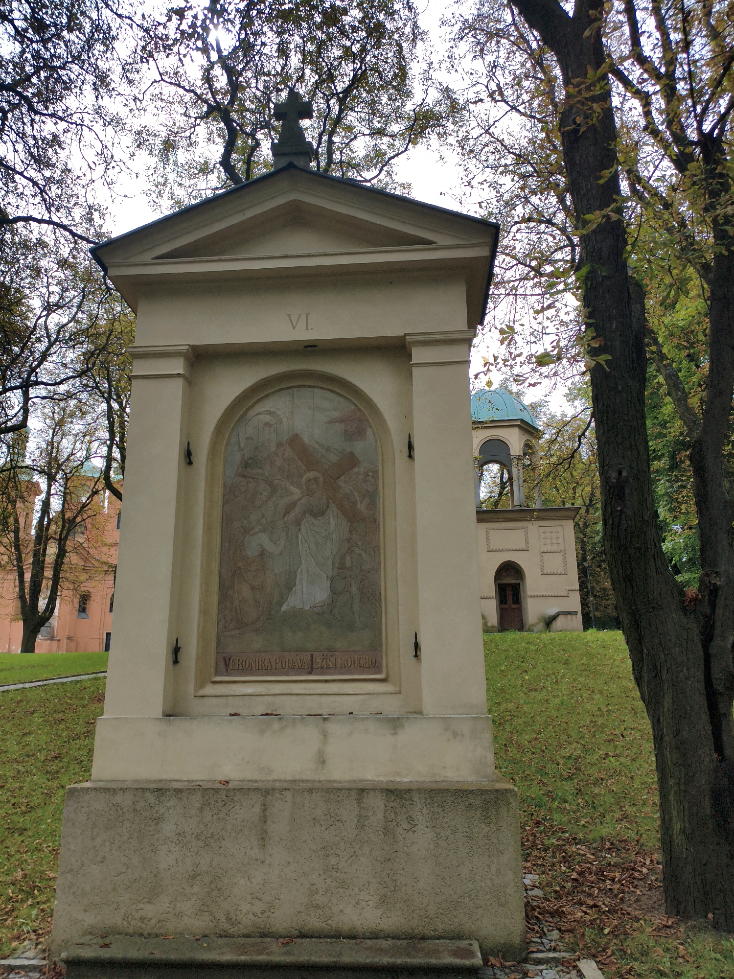 The image shows a devotional structure, likely a station of the cross, in an outdoor setting. The structure is made of stone and features a painting on its front panel, depicting a religious scene with multiple figures. Above the painting, the Roman numeral "VI" is visible. The surrounding environment is autumnal, with trees displaying sparse foliage and fallen leaves on the ground. In the background, there is a building with a light-colored facade and a turquoise dome, possibly a chapel or part of a larger complex. The overall impression is one of a serene, possibly historical, religious site within a park or garden. The devotional structure is a prominent element, drawing the viewer's attention to the painted scene and the inscription. The presence of trees and fallen leaves suggests a season of change and perhaps contemplation.