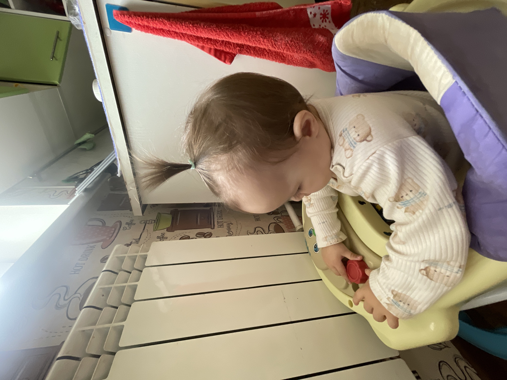 In Sharkan, Russia, a toddler with a high ponytail is seated in a baby activity center. The toddler is wearing a white long-sleeved shirt with a pattern of teddy bears. Their hands are engaged with the red buttons on the activity center. The background features a radiator against a wall with a coffee-themed wallpaper. A red towel hangs on the right side of the image. The lighting suggests an indoor environment, possibly a home.