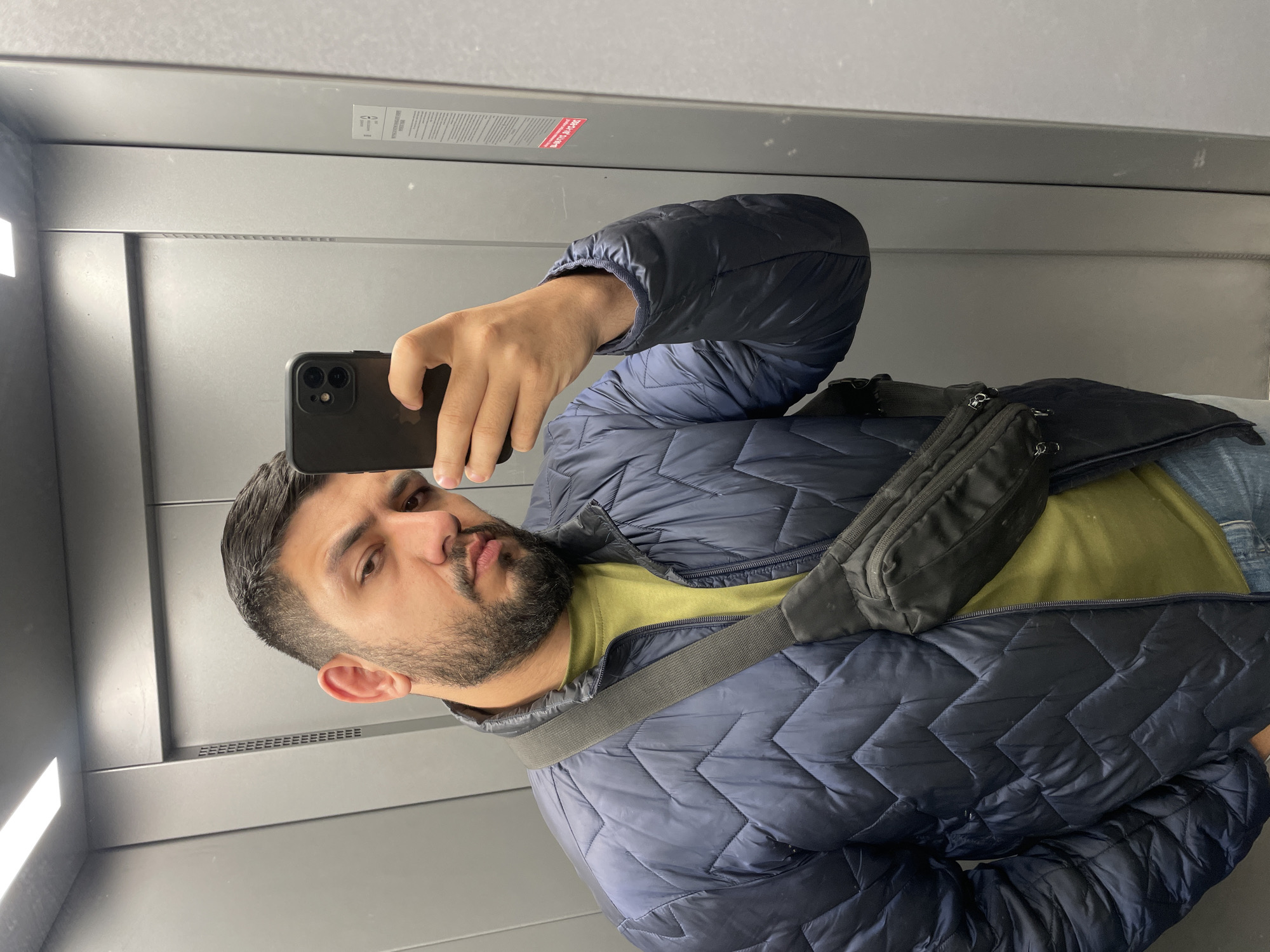 A man with a beard is taking a selfie in what appears to be an elevator. He is wearing a blue quilted jacket over a green shirt, with a black crossbody bag. The elevator walls are gray. The location is unknown, but based on the prompt, it is presumed to be in Krasnoyarsk, Russia.