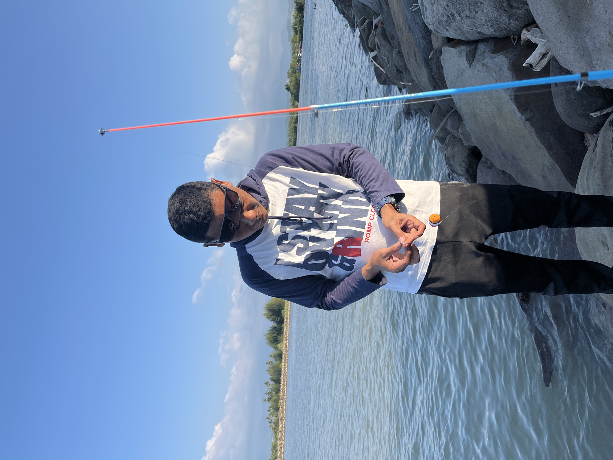 A man stands on a rocky shoreline in Reudeuep, Indonesia, preparing to fish. He is wearing sunglasses and a long-sleeved hooded shirt with a graphic print and dark blue sleeves, paired with black pants. In his hands, he holds a fishing line with a small orange bobber. A blue fishing rod with a red tip stands to his right. The background shows a calm body of water, a distant treeline with palm trees, and a clear blue sky with scattered clouds. The rocks he is standing on are large and grey.