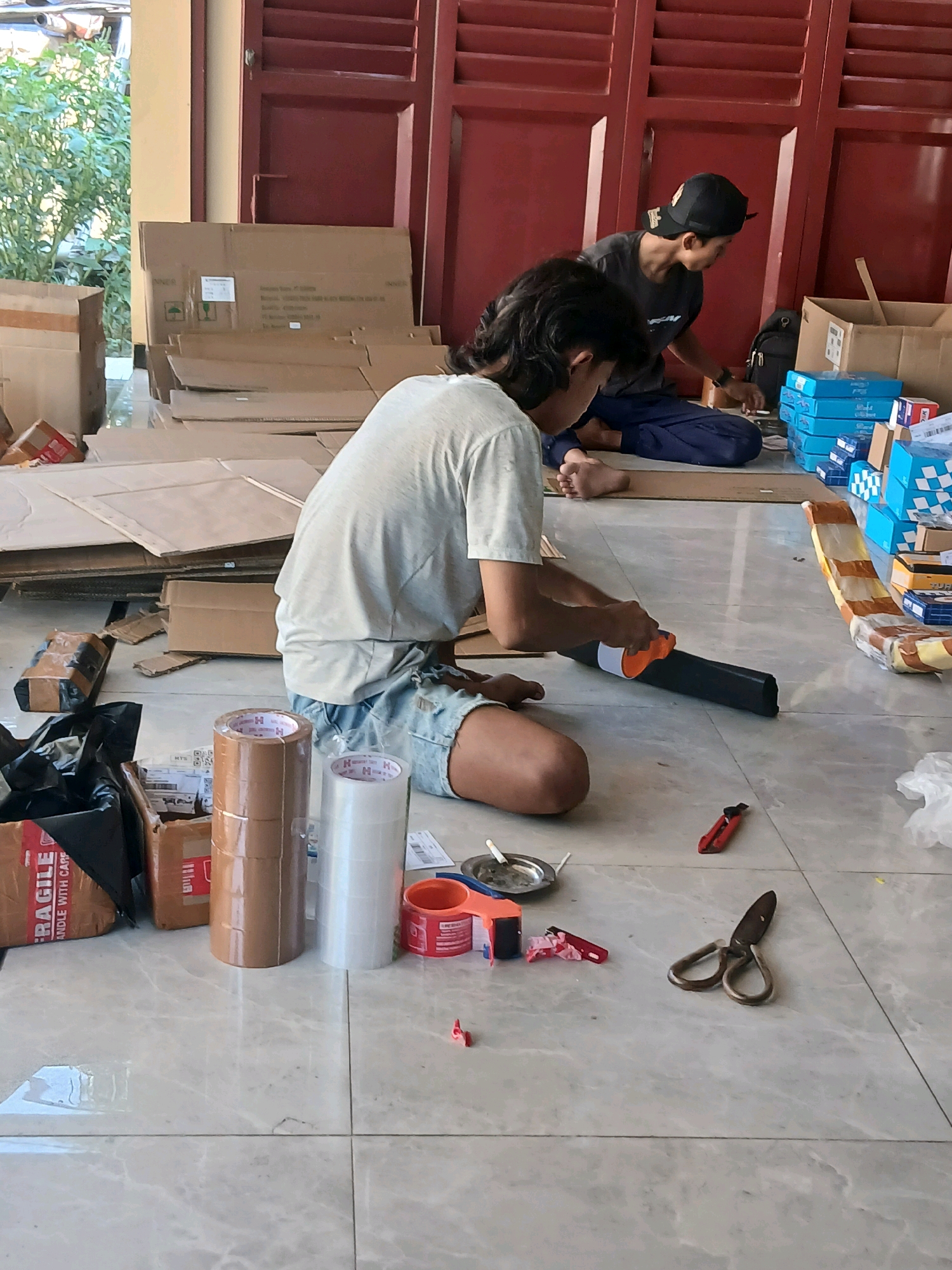 In the village of Jepangpakis, Indonesia, two individuals are observed engaged in packing activities. The scene is set on a tiled floor, with numerous cardboard boxes, both assembled and disassembled, surrounding the subjects. One individual, with long dark hair and wearing a light-colored t-shirt and denim shorts, is seated on the floor and actively using a tape dispenser to seal a cardboard tube. The other individual, wearing a dark t-shirt and a black baseball cap, is also seated and appears to be sorting or organizing items, with a stack of blue boxes visible to their right. Various packing materials are scattered on the floor, including rolls of packing tape, a box cutter, a pair of scissors, and what appears to be an ashtray with a cigarette butt. The environment suggests an informal, possibly domestic or small business setting, with large red doors and green foliage visible in the background.