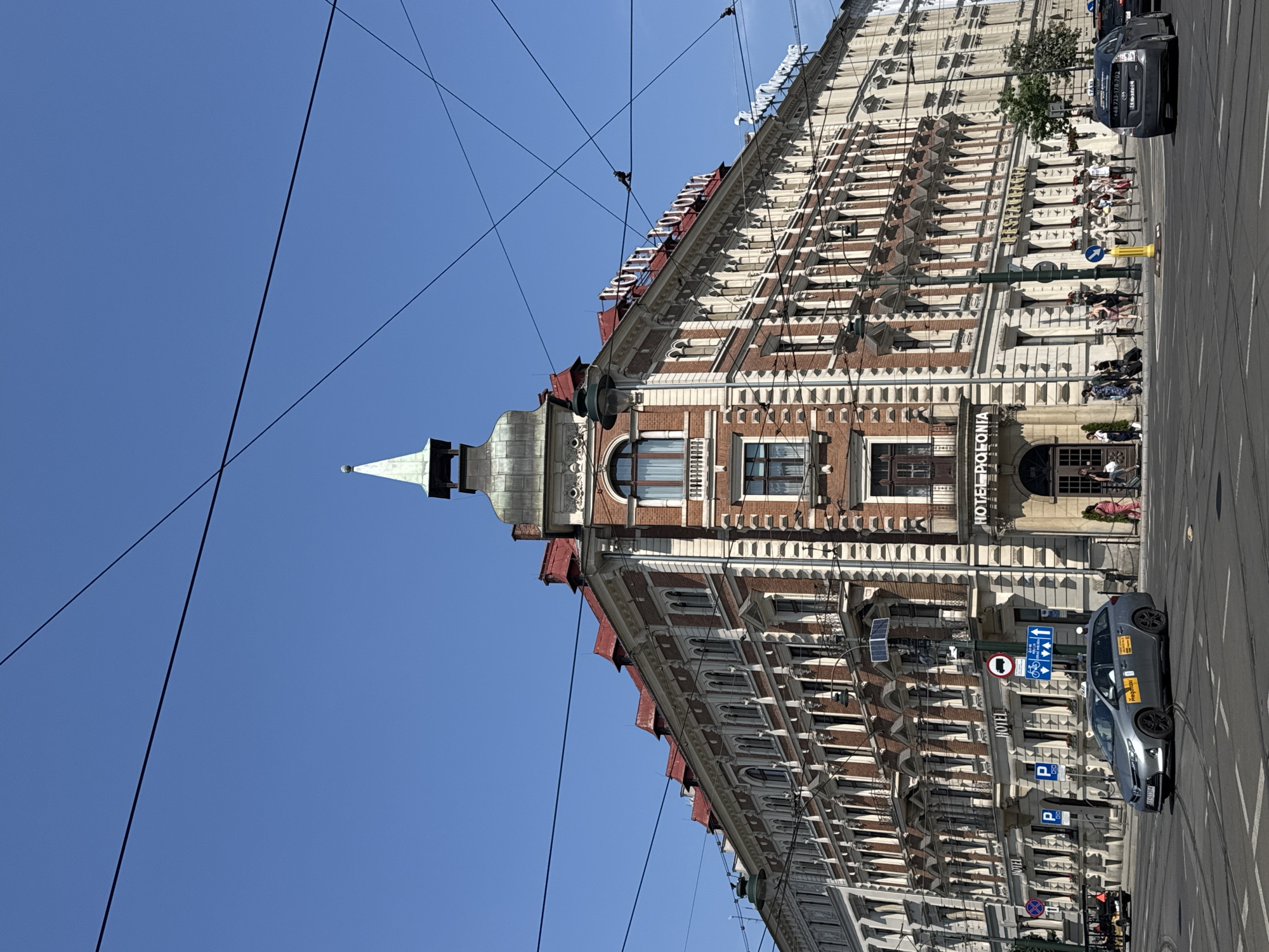 The image shows a street view of the Hotel Polonia in Krakow, Poland, on a clear, sunny day. The hotel is a large, ornate building with red tiled roofs and red brick facades, featuring multiple floors of arched windows and decorative architectural details. A prominent sign on the building reads "HOTEL POLONIA".
In the foreground, a silver Toyota Prius taxi is driving on the street. The taxi has "MegaTaxu" written on its side. Several other cars are visible on the road, as well as pedestrians walking on the sidewalk in front of the hotel. There are traffic lights, street signs, and electrical wires crisscrossing the sky above the street. The street is paved and appears to be a busy intersection. The overall environment suggests an urban setting with active street life.