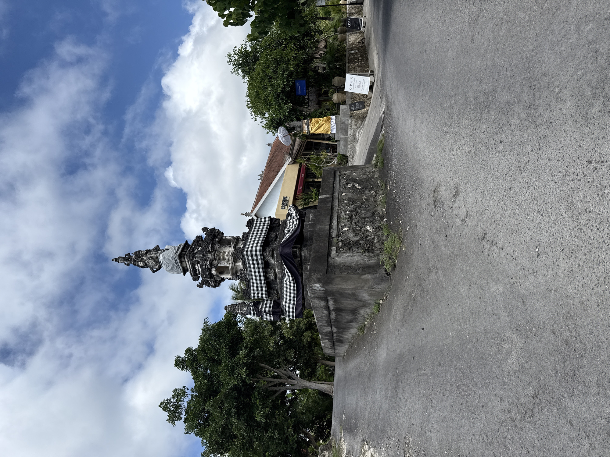 The image shows a Balinese shrine located on the side of a road in Pejukutan, Indonesia. The shrine is made of carved stone, decorated with a black and white checkered cloth. It stands on a concrete base. A paved road runs alongside the shrine. In the background, trees and buildings are visible, along with signage. The sky is blue with scattered clouds. The shot appears to be taken from a low angle.