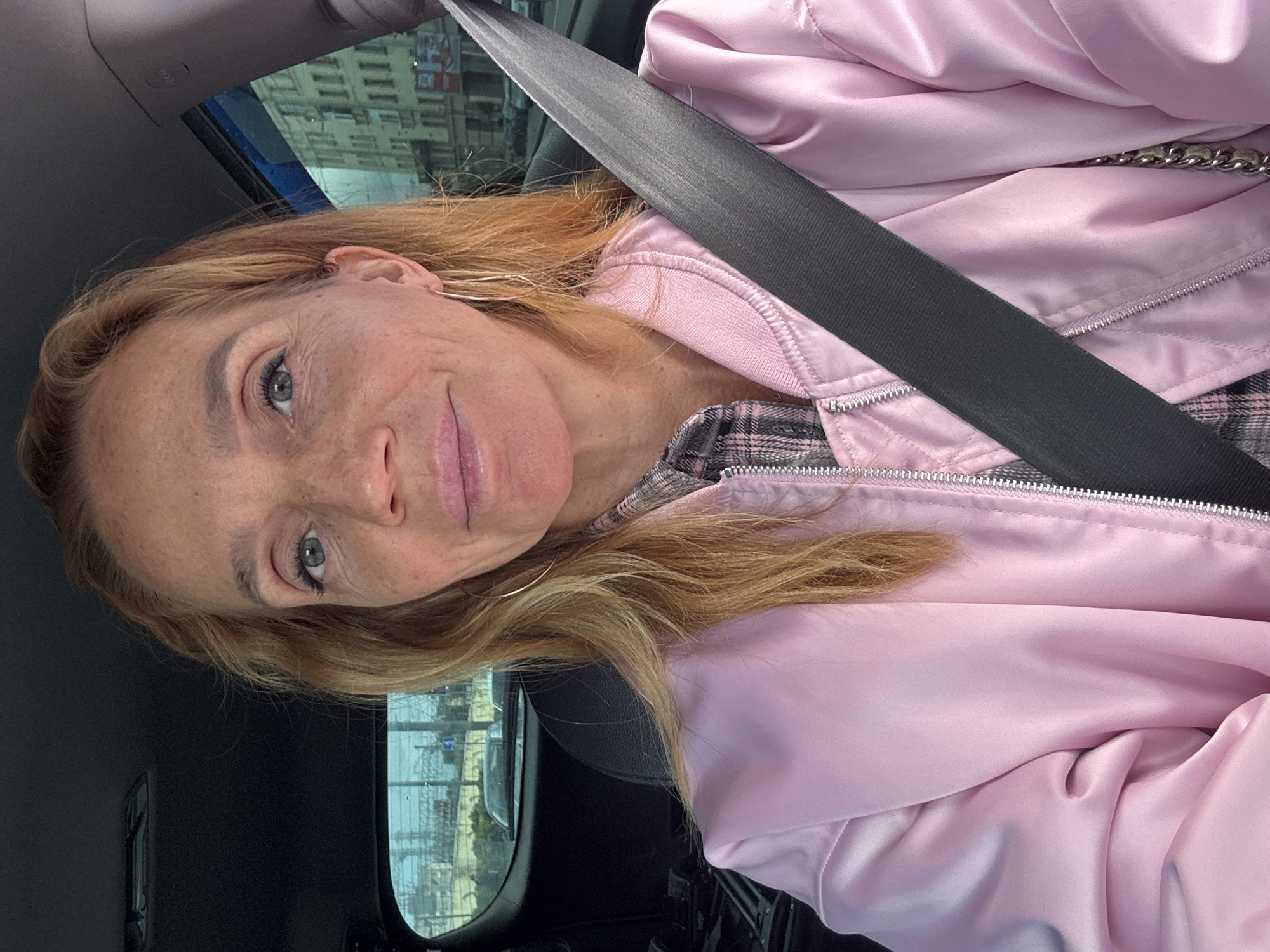 A woman with blonde hair and freckles is sitting in the driver's seat of a car. She is wearing a pink bomber jacket over a plaid shirt and a black seatbelt is across her chest. Her left hand is visible near her lap and she is wearing a large hoop earring in her left ear. The car appears to be moving on a street in Saint Petersburg, Russia, as evidenced by the architecture visible through the car's windows. Specifically, a multi-story building with many windows and balconies, typical of St. Petersburg's historic districts, can be seen. The sky is overcast.
