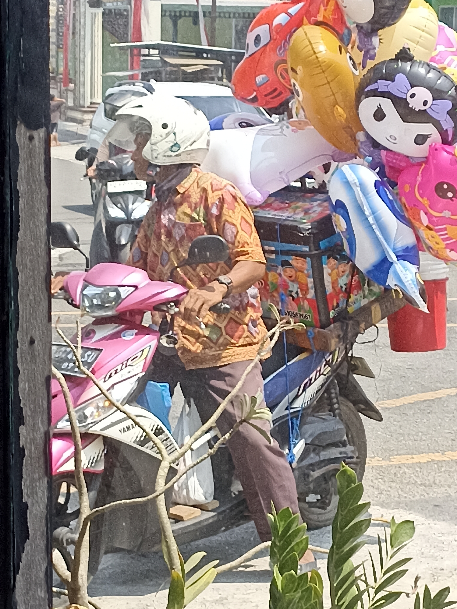 In Pulisen, Indonesia, a man wearing a white helmet and a patterned shirt rides a pink and white scooter. He is carrying a large assortment of colorful balloons, including a red car-shaped balloon, a yellow balloon shaped like a fish, and a black and white character balloon with a skull bow. The balloons are attached to a box mounted on the back of the scooter, which is decorated with pictures of children. The scooter is parked on a street with other vehicles and buildings in the background.