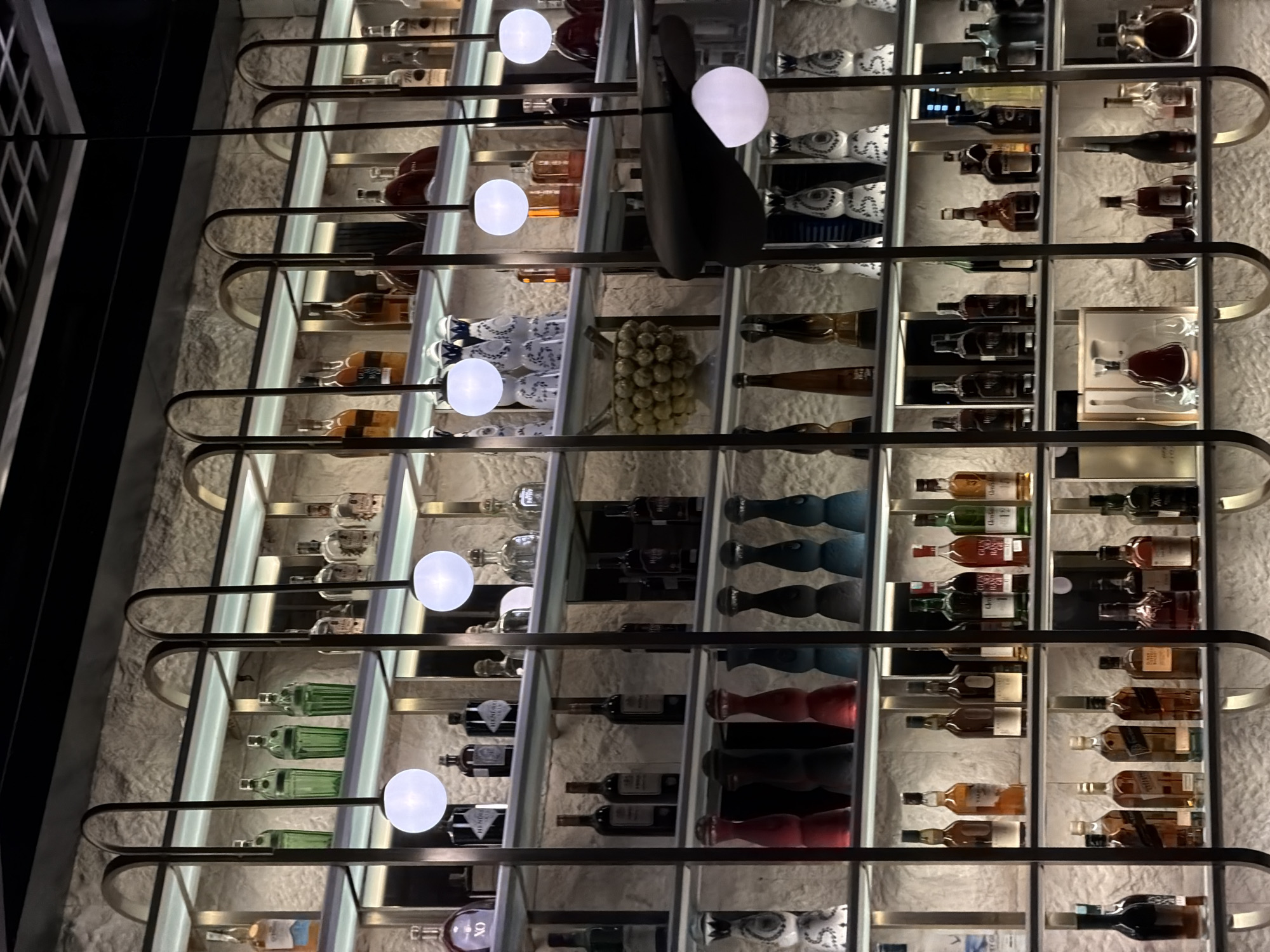 The image displays a vertically oriented, elaborate shelving unit filled with a diverse collection of alcoholic beverages, set against a rough, light-colored stone or stucco wall. The unit features multiple tiers supported by a modern, curved metal framework, and each shelf is illuminated from below, casting a warm glow on the bottles. Several spherical white lights are integrated into the metal structure, further brightening the display.
The shelves hold numerous bottles of spirits, including recognizable brands of whisky such as Glenfiddich, Talisker, and Johnnie Walker Gold Label Reserve. There are also distinctive bottles of Hennessy cognac, including one marked "XO" presented in a box with glassware. Other bottles of clear spirits, green-tinted glass bottles, and wine bottles are visible. Interspersed among the liquor bottles are decorative objects, such as white and blue patterned ceramic vessels, and groups of blue/teal and red shaped glass objects. A cluster of small, pale yellow spherical items is also displayed on one shelf. A large, dark, abstract-shaped object hangs from an upper shelf.
The lighting suggests an evening or night setting within an indoor commercial establishment, such as a bar or a high-end restaurant lounge. No people or specific activities are visible, focusing solely on the ornate display. On the far left, a dark area with a white grid pattern suggests an adjacent window or decorative grille.