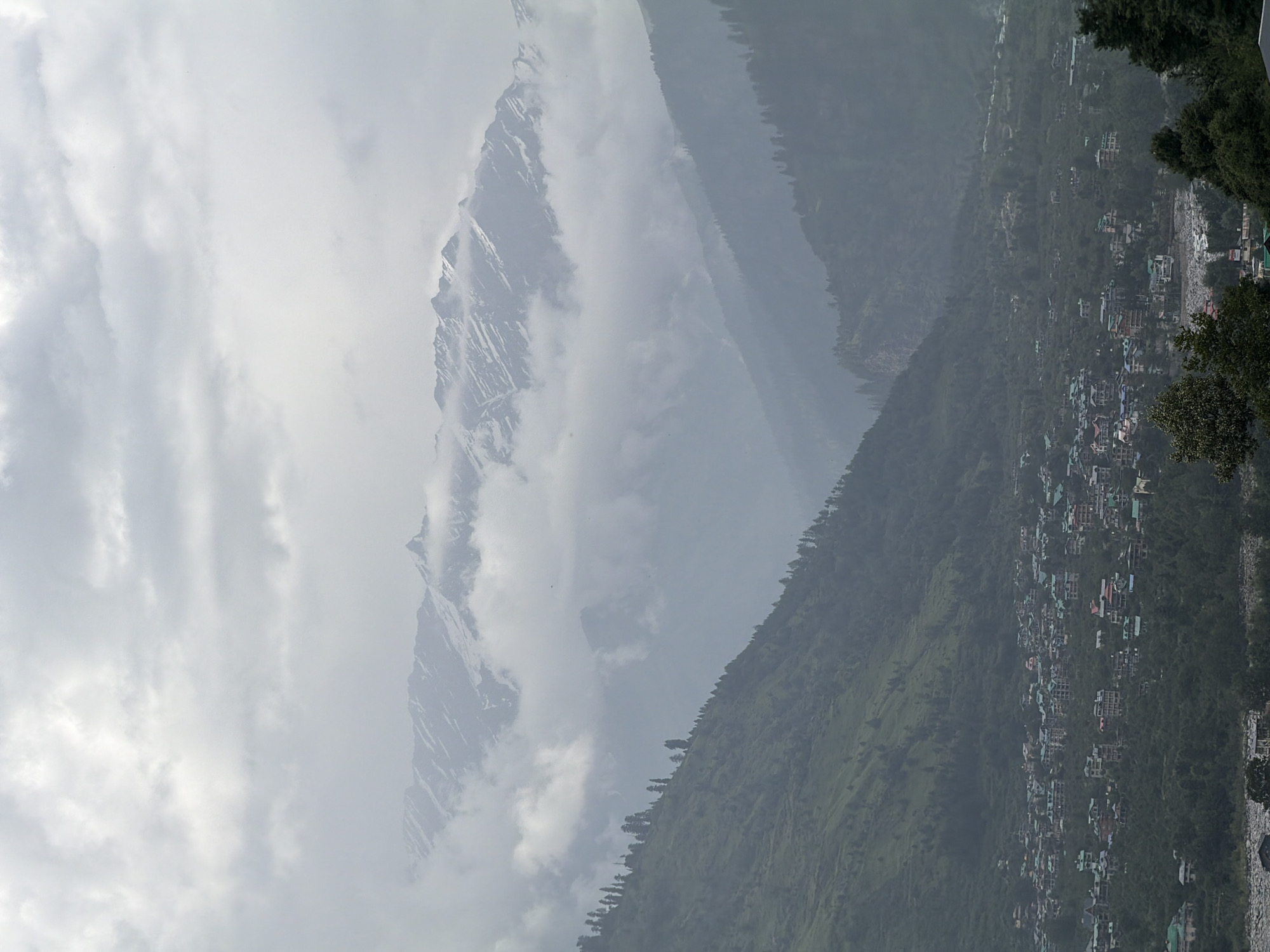 This image captures a panoramic view of the town of Vashisht, India, nestled within a valley at the foothills of snow-capped mountains. The scene is dominated by a hazy, overcast sky, with layers of clouds obscuring the upper reaches of the distant, rugged mountain range. Patches of snow are visible on the mountain peaks, contrasting with the darker, mist-shrouded slopes.
In the mid-ground, the steep, verdant slopes of the valley are densely covered with trees, creating a textured, natural landscape. A small town, identified as Vashisht, is spread out across the lower slopes and valley floor. The buildings are predominantly small to medium-sized structures, many with colorful roofs, indicative of a residential and possibly tourist area. The architecture appears to be a mix of traditional and modern styles, with clusters of houses and hotels.
The foreground features the lower slopes of the valley, also covered in dense vegetation and trees. A river or stream can be seen in the lower part of the image, reflecting the overcast sky. There are no visible people or animals in the frame. The overall atmosphere is one of a quiet, mountainous town under a moody, cloudy sky, emphasizing the natural beauty and the human settlement integrated within it.