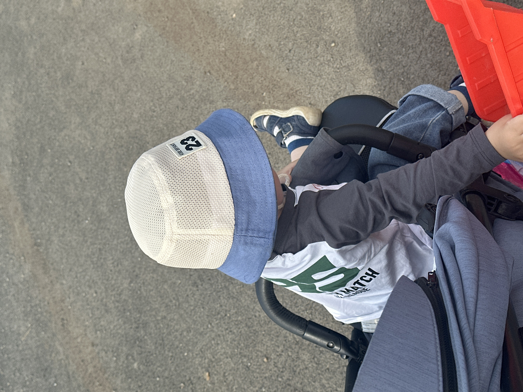 A child wearing a hat with a "23" patch and sandals is seated in a stroller with a red container in the background. The stroller is positioned on a gray surface, potentially a sidewalk. The child wears a long-sleeved gray shirt over a white T-shirt with text that includes the word "MATCH."