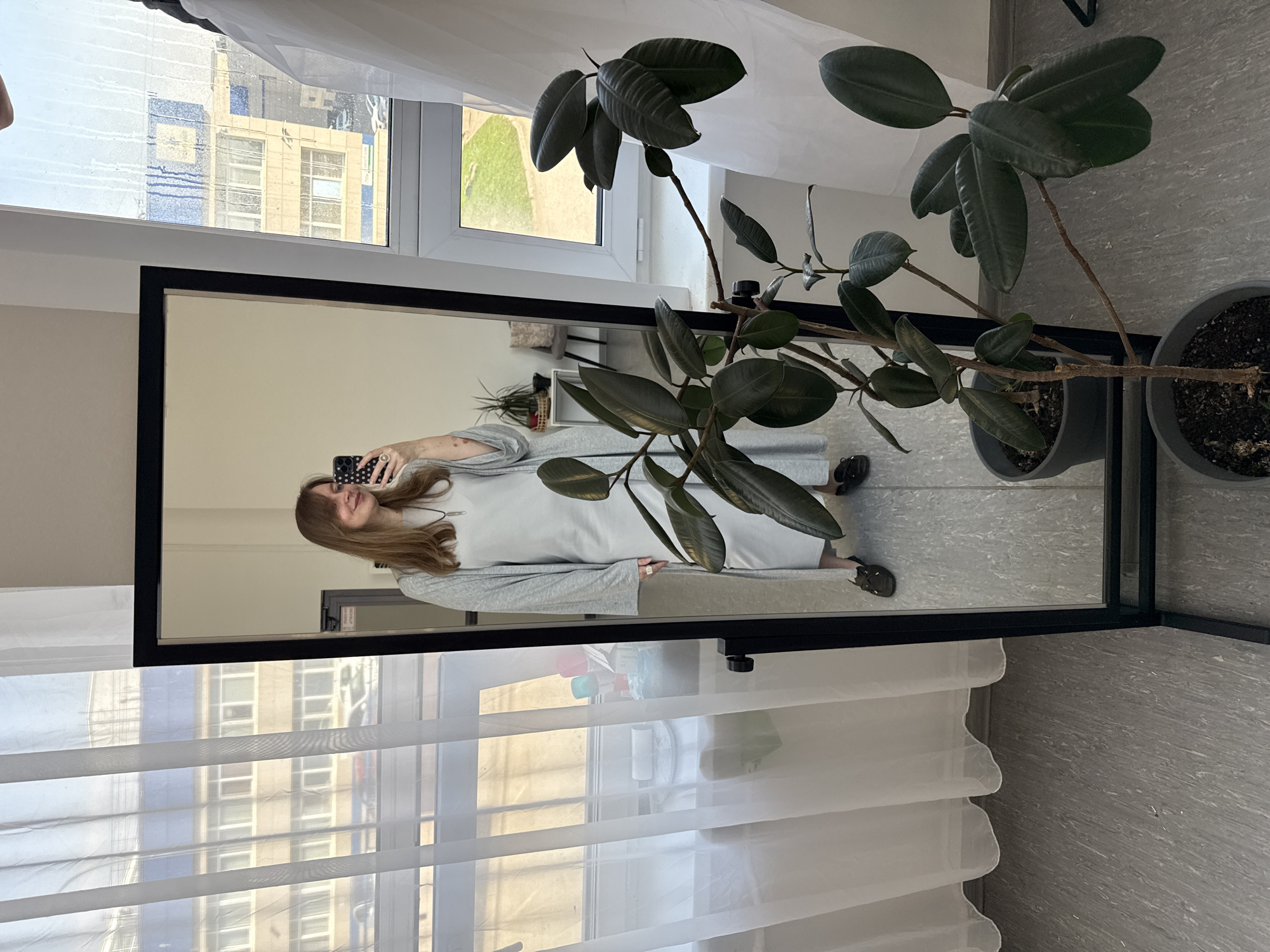 A woman is reflected in a full-length mirror in an indoor setting. She is holding a phone and appearing to take a photo of herself in the mirror. She is wearing a grey dress and light grey cardigan. The mirror is standing near a window covered with semi-transparent white curtains. Through the curtains, buildings are visible, suggesting an urban environment. A large potted rubber plant stands to the right of the mirror, partially obscuring the view of the woman. The floor is tiled with light grey tiles. The location is Izhevsk, Russia, based on the appearance of the buildings seen through the window.