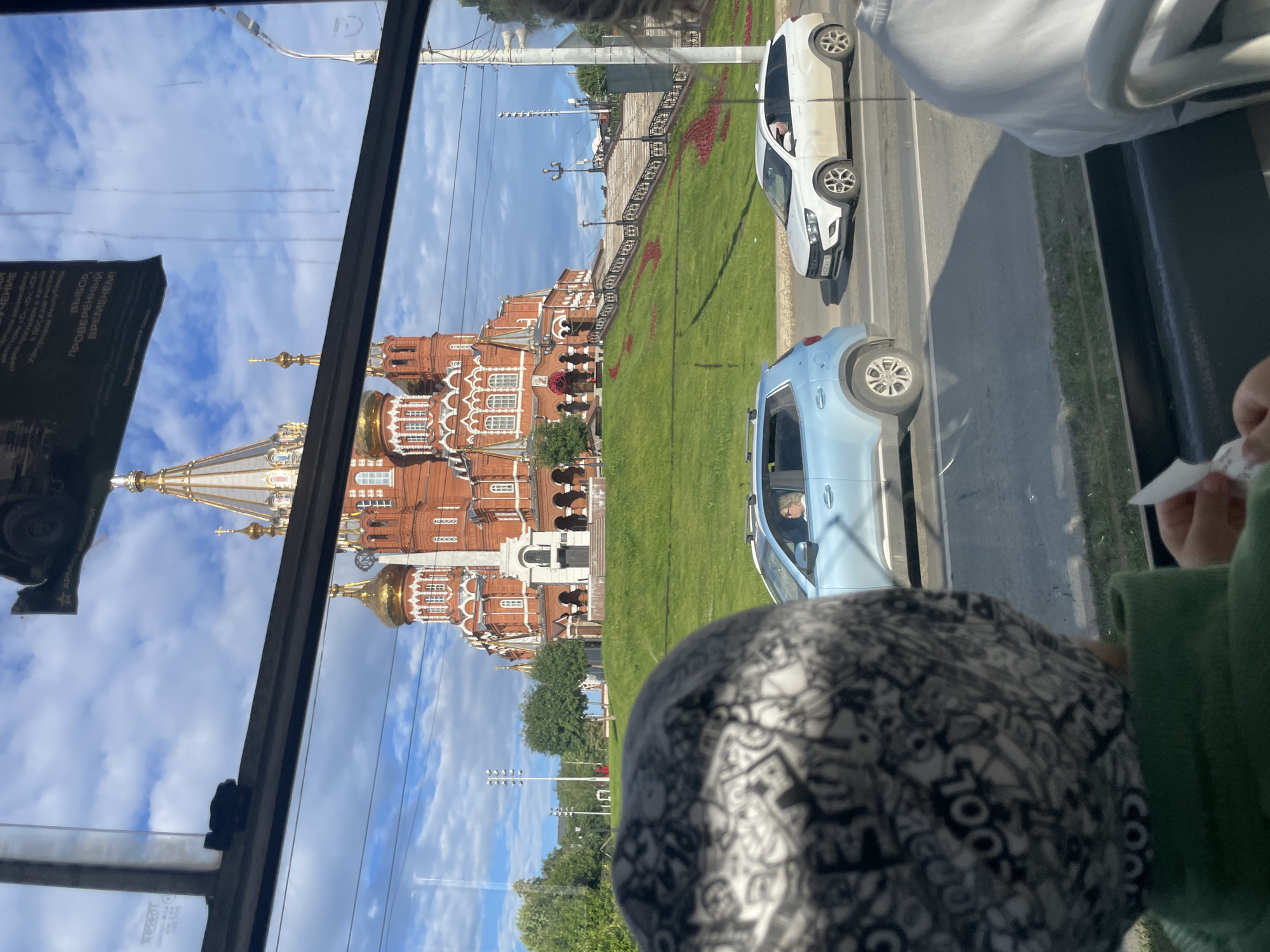 The image, taken from inside a vehicle, shows a view of a large, ornate red brick building with golden domes, likely a cathedral or church, under a partly cloudy sky. The building is adorned with intricate white detailing and appears to be a prominent landmark. In front of the building is a lush green lawn with red floral arrangements, forming a decorative pattern. Two cars, a white sedan and a light blue SUV, are visible on a road adjacent to the lawn. The perspective suggests the viewer is looking out of a window, with a portion of a person's head, wearing a patterned hat, and a hand holding a white slip of paper in the foreground. Reflected on the glass, some text is visible, including "ЧЕНИЕ", "ВЫБОР, ПРОВЕРЕННЫЙ ВРЕМЕНЕМ!", and what appear to be fragments of Russian phrases. The scene suggests a daytime outing in a city, possibly Izhevsk, Russia, given the textual cues. The overall impression is one of sightseeing or traveling, with the subjects observing the historical architecture and cityscape. The light suggests it is daytime.
