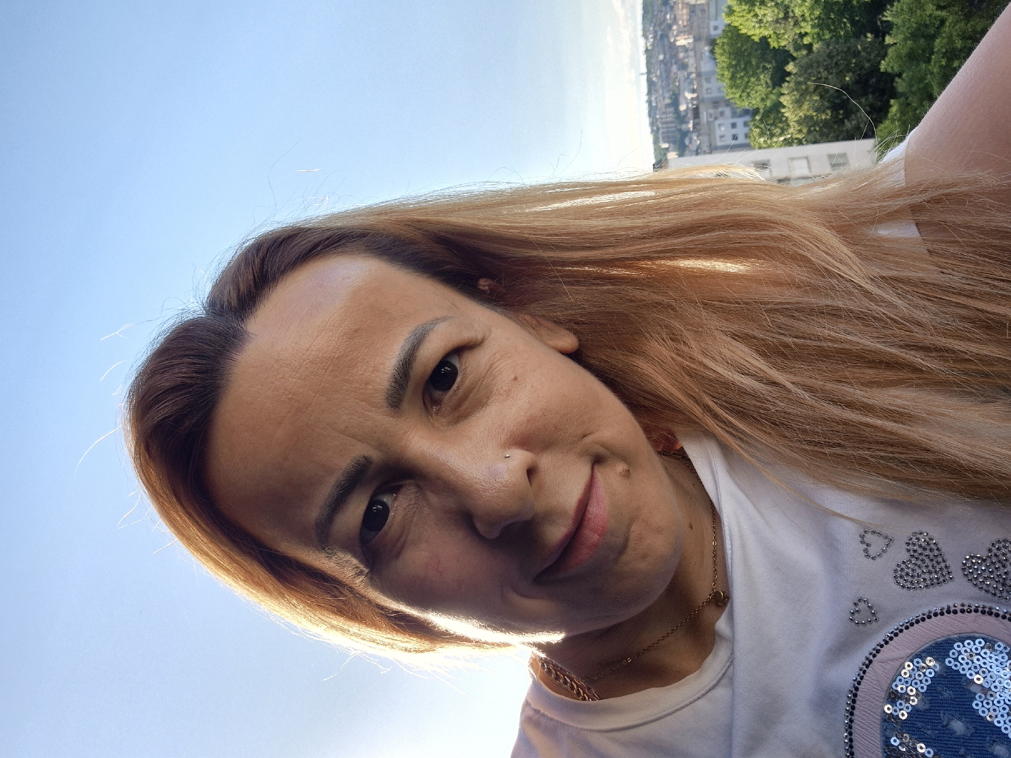 A close-up selfie of a person with auburn hair and fair skin, looking towards the camera with a slight smile. The person is wearing a white t-shirt with a chain necklace and a small pendant. They have a nose ring and some makeup on. The background shows a clear blue sky, buildings, and green trees, suggesting an urban outdoor setting. The lighting indicates it is daytime, likely late afternoon due to the angle of the sun.