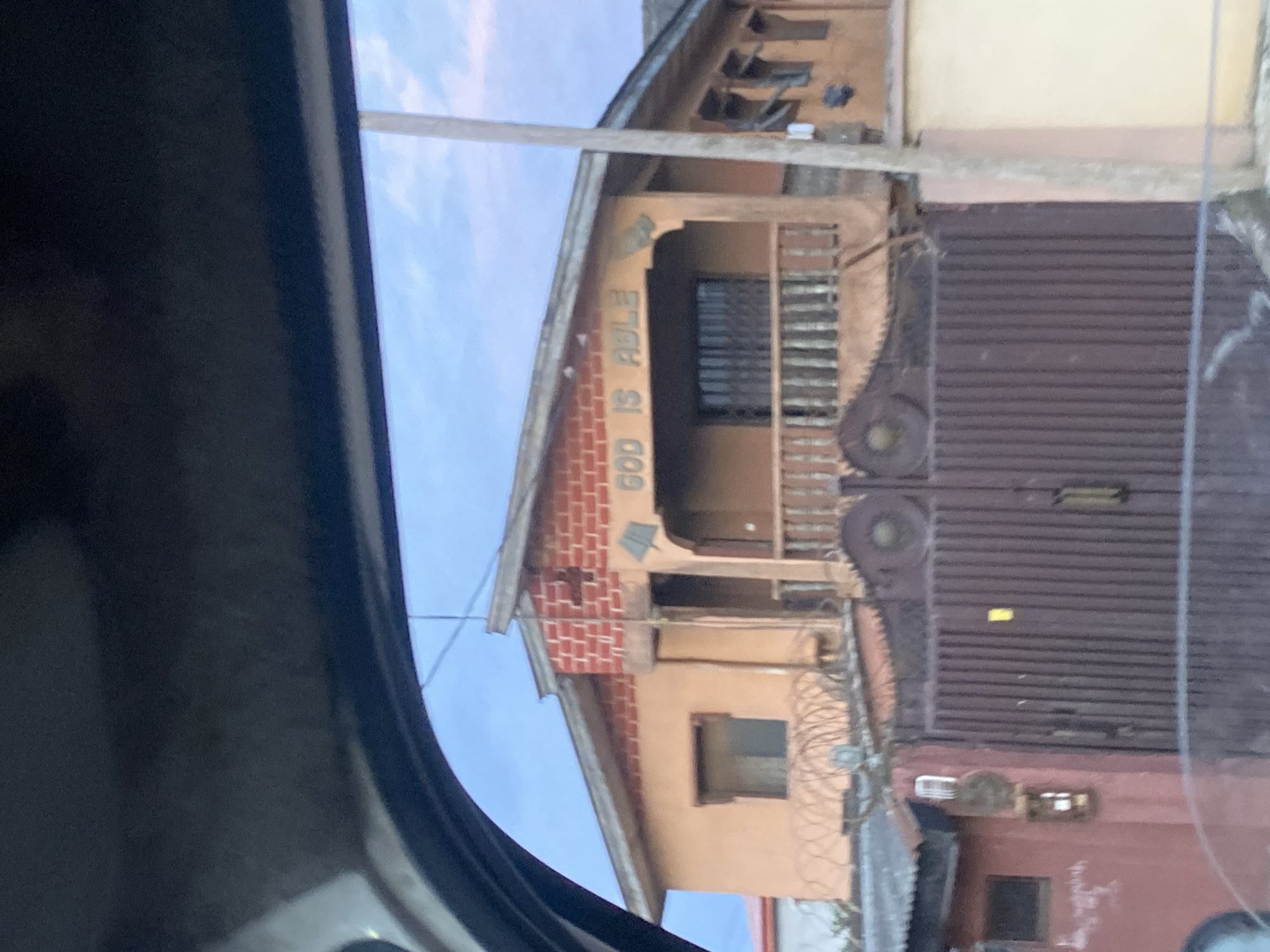 The image depicts a building facade in Ikorodu, Nigeria, viewed from a slightly elevated perspective, likely from within a vehicle. The building is painted in a peach-like color and features a prominent brick section on the upper front. Above the balcony, the words "GOD IS ABLE" are inscribed in raised lettering. The balcony itself is enclosed by a railing and has barred windows. In front of the building, there is a large, corrugated metal gate, dark brown in color, with decorative elements. Barbed wire is visible along the top of a lower wall or fence in front of the gate. The sky above is overcast, with hints of blue and scattered clouds, suggesting it is either early morning or late afternoon. The scene appears to be a residential or mixed-use property within a built-up urban environment.