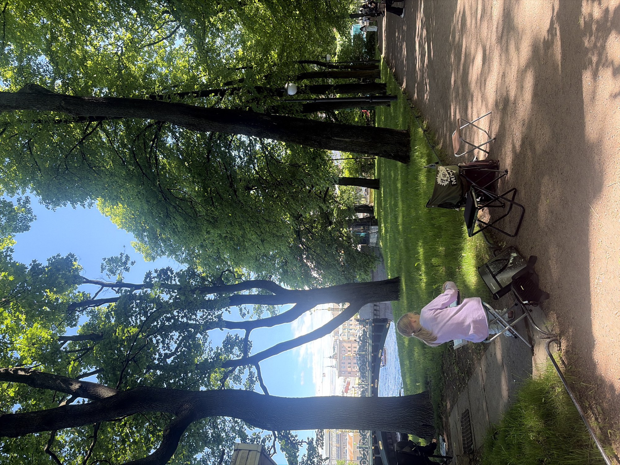 The image depicts a bright, sunny day in a park-like setting in Saint Petersburg, Russia, likely along the Moika River embankment near Mikhailovsky Garden, looking towards the Malaia Konyushennaya Bridge and the cityscape beyond.
In the foreground, a young fair-haired girl, with her hair tied in a ponytail, sits on a small silver folding stool, facing away from the viewer. She wears a light purple long-sleeved shirt and light-colored pants. She is engaged in an artistic activity, appearing to draw or paint on a small white surface held on her lap or a portable easel. To her left, a black case or bag rests on the ground. A black folding stool, an empty lighter-colored folding stool, and a dark green canvas bag with white text, including "MIPA" and other characters, are positioned on the reddish-brown dirt path to her right. A dark cable or hose runs along the edge of the path near her.
The path, casting dappled tree shadows, stretches into the background on the right, lined by several mature trees with dark trunks and lush green foliage. Street lamps with white globes are visible along the path. In the midground, a wide, green grassy area separates the path from a body of water, likely the Moika River. Two large tree trunks frame a view across the water.
Across the water, a green metal arch bridge with ornate railings and lamp posts spans the river. Beyond the bridge, a vibrant city skyline features historical European-style buildings with varied rooflines, some topped with decorative elements and small domes, under a clear blue sky with scattered white clouds. A small boat is visible on the river beneath the bridge. Further along the path in the distance, blurred figures of other people are discernible.