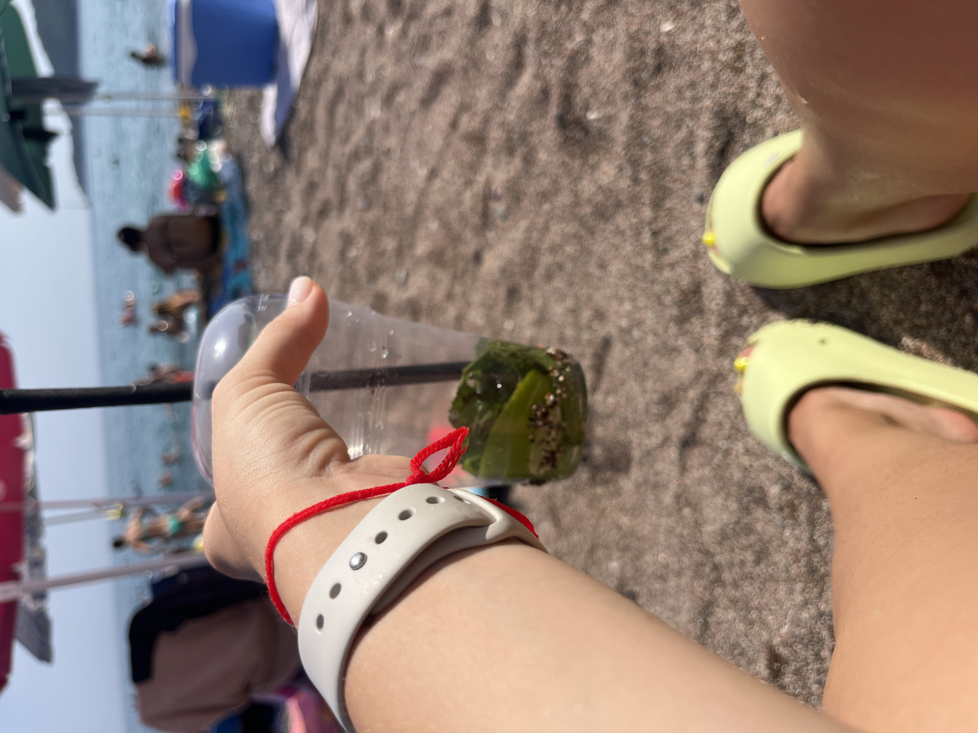 A first-person perspective shot of someone holding a clear plastic cup filled with ice, lime slices, and possibly mint leaves, on a beach in Boreti, Montenegro. The cup is held in the right hand, which has a white fitness tracker on the wrist and a red string bracelet. The background is blurred, showing the sea with people swimming, beach umbrellas, and beachgoers relaxing on the sand. The sand is granular and light brown. The person is wearing bright yellow slides. The scene suggests a relaxed, sunny day at the beach.