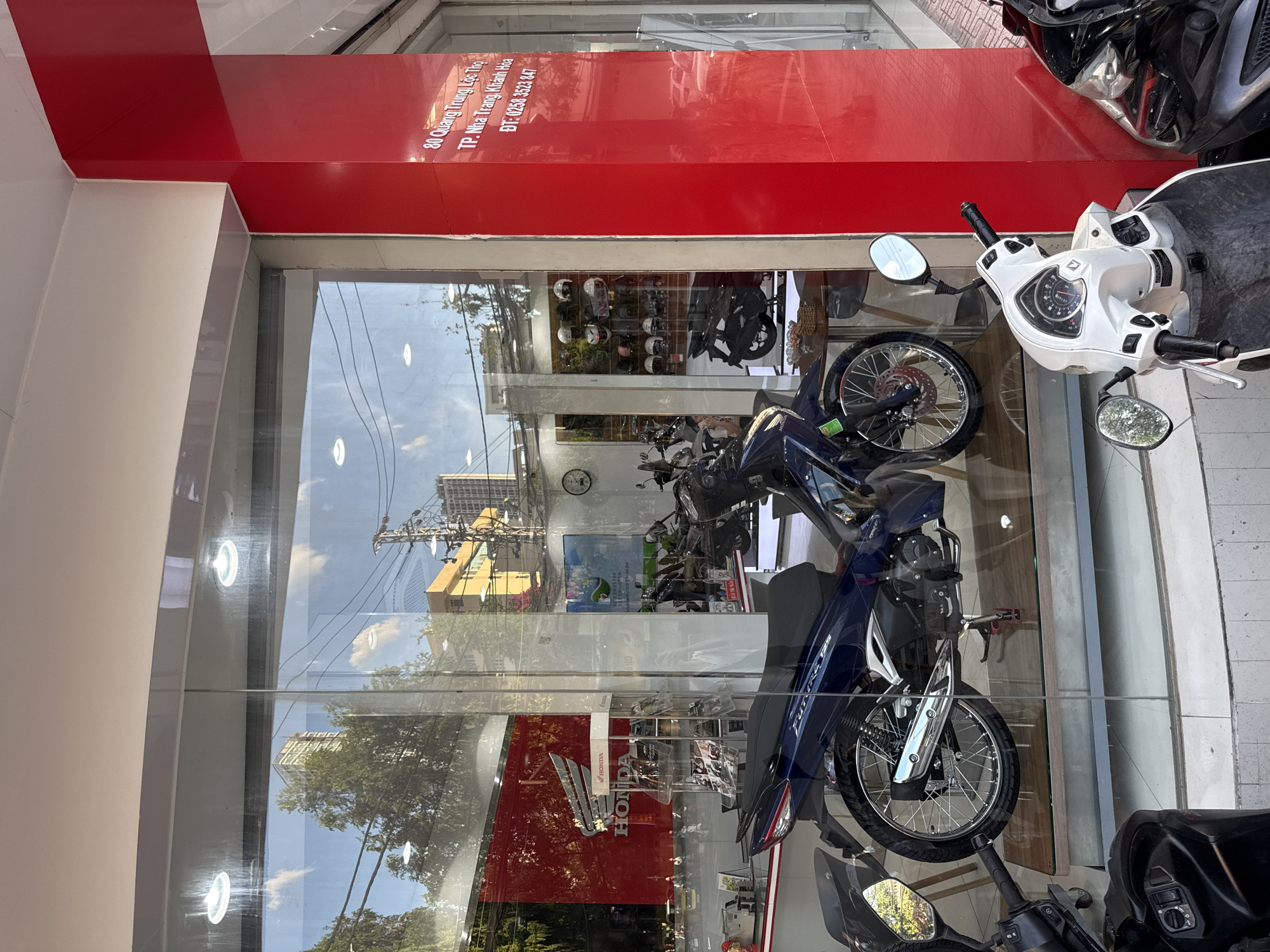 The image shows the exterior of a Honda motorcycle dealership in Nha Trang, Vietnam, with its motorcycles displayed behind a large glass window. The dealership is located at 80 Quang Trung Street, Loc Tho Ward. A blue Honda motorcycle is prominently displayed in the window, as well as a white scooter and a black scooter outside the window. The interior of the dealership is visible, with helmets on display and other motorcycles. The exterior of the building is painted red and white, and the street is visible through the glass. The sky is clear with some clouds.