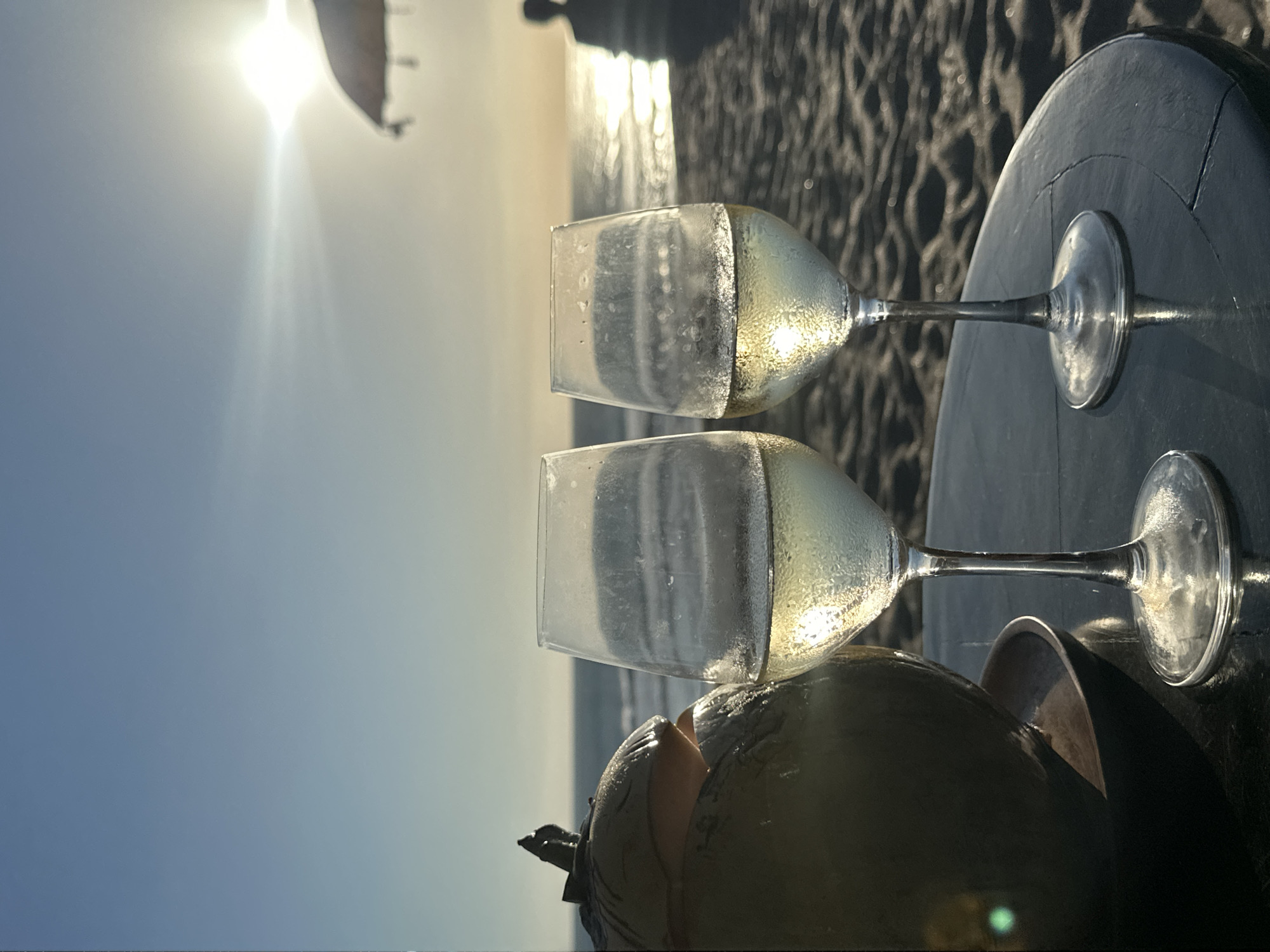 An outdoor, eye-level shot captures two wine glasses and a coconut drink on a dark, circular table against a vibrant beach sunset or sunrise. The image is oriented vertically, showing the horizon line on the right side.
In the foreground, two stemmed wine glasses, frosty from cold liquid, are partially filled with a light, possibly white wine or a pale cocktail. Below them, a green coconut is visible, cut open at the top, likely serving as a natural drinking vessel. These items rest on a dark, textured round table, with reflections of the bright light playing on its surface.
The background is dominated by the dazzling sun, emitting a strong lens flare and illuminating the scene with a warm, golden glow. The sun's light reflects brightly off a calm body of water, likely the ocean, creating a shimmering path. Beyond the water, the texture of sand or a beach is visible. A dark, blurred object, possibly a part of a boat's sail or a kite, can be vaguely discerned near the sun. The sky transitions from a soft blue at the bottom left to a brighter, warmer hue closer to the sun.
The overall scene conveys a serene and relaxing moment, perhaps a beachside toast during a sunset or sunrise in a tropical location like North Kuta, Indonesia. There is no visible text in the image.