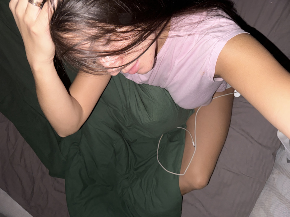 A female individual is depicted lying on a bed in an indoor setting, presumably a residence in Troitsk, Russia. The bed is furnished with dark grey sheets and a dark green duvet or blanket, which partially covers the lower body of the individual. The female has long, dark hair that partially obscures her face, and her eyes appear closed or mostly closed. She is wearing a light pink t-shirt. Her right arm is raised, with the hand resting on the back of her head, displaying a ring on her right ring finger. White wired earbuds are visible, with the cable extending from near her torso, looping across her left thigh, and disappearing off-frame. The scene suggests a state of rest or sleep.