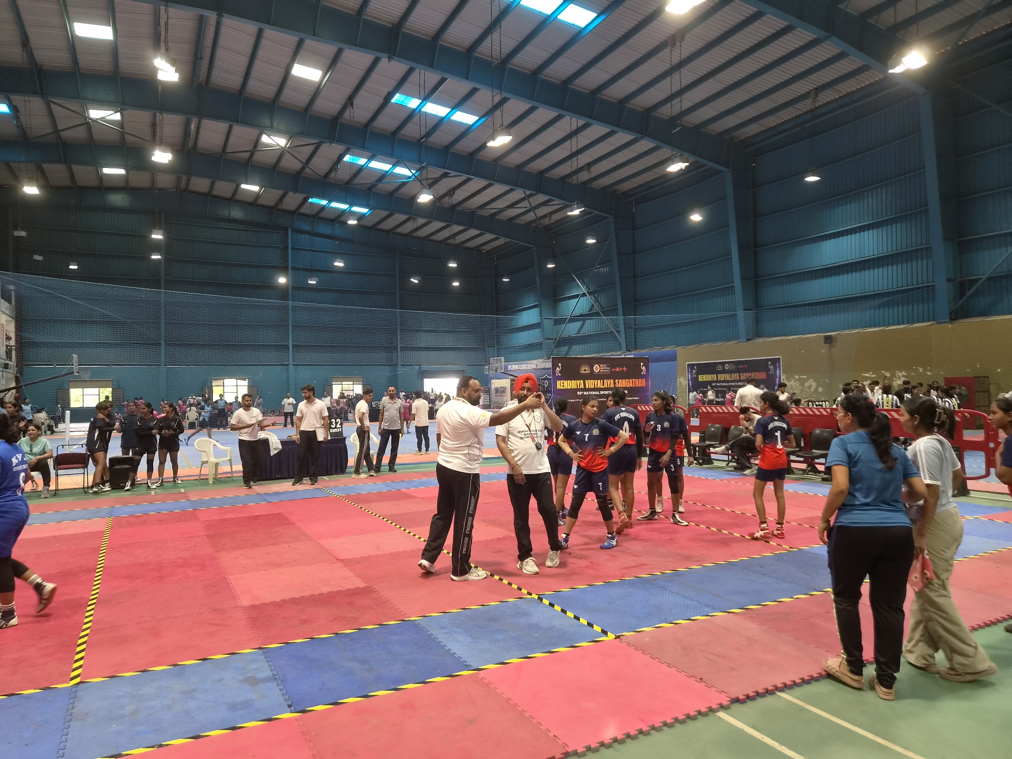 An indoor sports hall in Phagwara, India, hosts a kabaddi match. Red and blue mats delineate the playing field, marked with yellow and black lines. Several individuals, appearing to be participants and officials, are present on the court and surrounding areas. Some are dressed in athletic uniforms, while others wear casual attire. In the background, a large banner displays "KENDRIYA VIDYALAYA SANGATHAN 59TH NATIONAL SPORTS MEET." The hall features a high, corrugated metal roof with numerous lights and skylights. Spectators can be seen in the bleachers in the far background. The overall scene depicts an organized sporting event within a modern gymnasium.