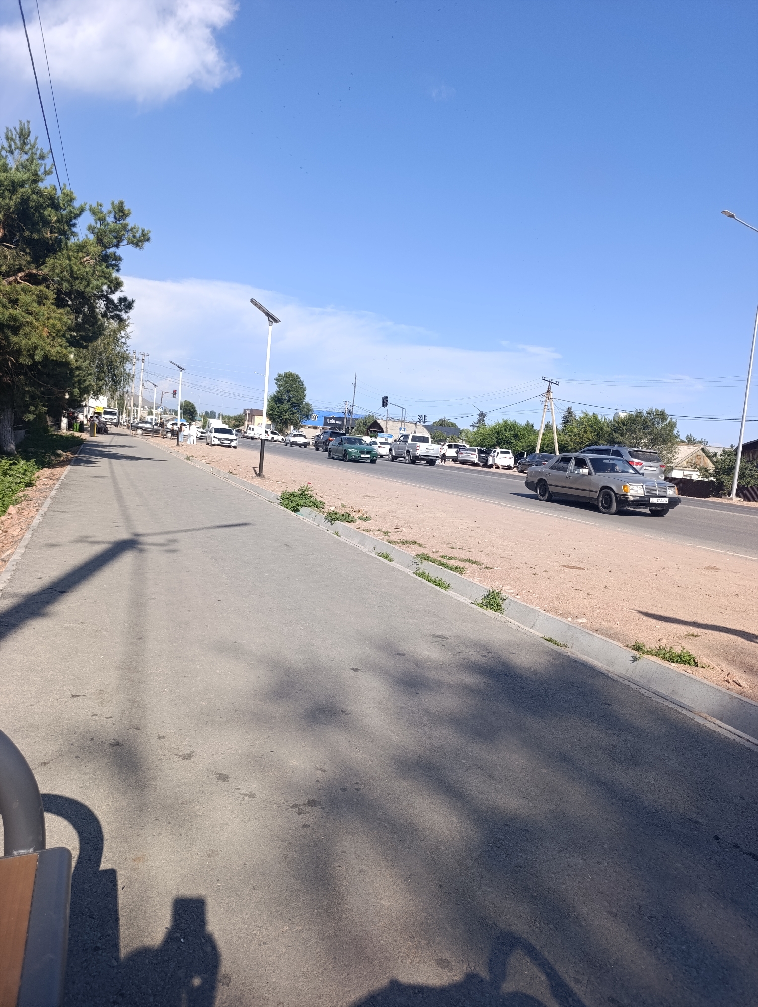 A sunny daytime scene on a street in Bosteri, Kyrgyzstan, shows a paved pedestrian walkway alongside a two-lane road. Trees line the left side of the walkway, casting shadows on the pavement. Several vehicles are visible on the road, including a silver Mercedes-Benz sedan in the foreground, a green sedan, a white van, and various other cars and trucks further down the road. Streetlights are intermittently spaced along the road. The sky is blue with scattered white clouds. The environment appears to be a mixture of urban and residential, with buildings visible in the background.