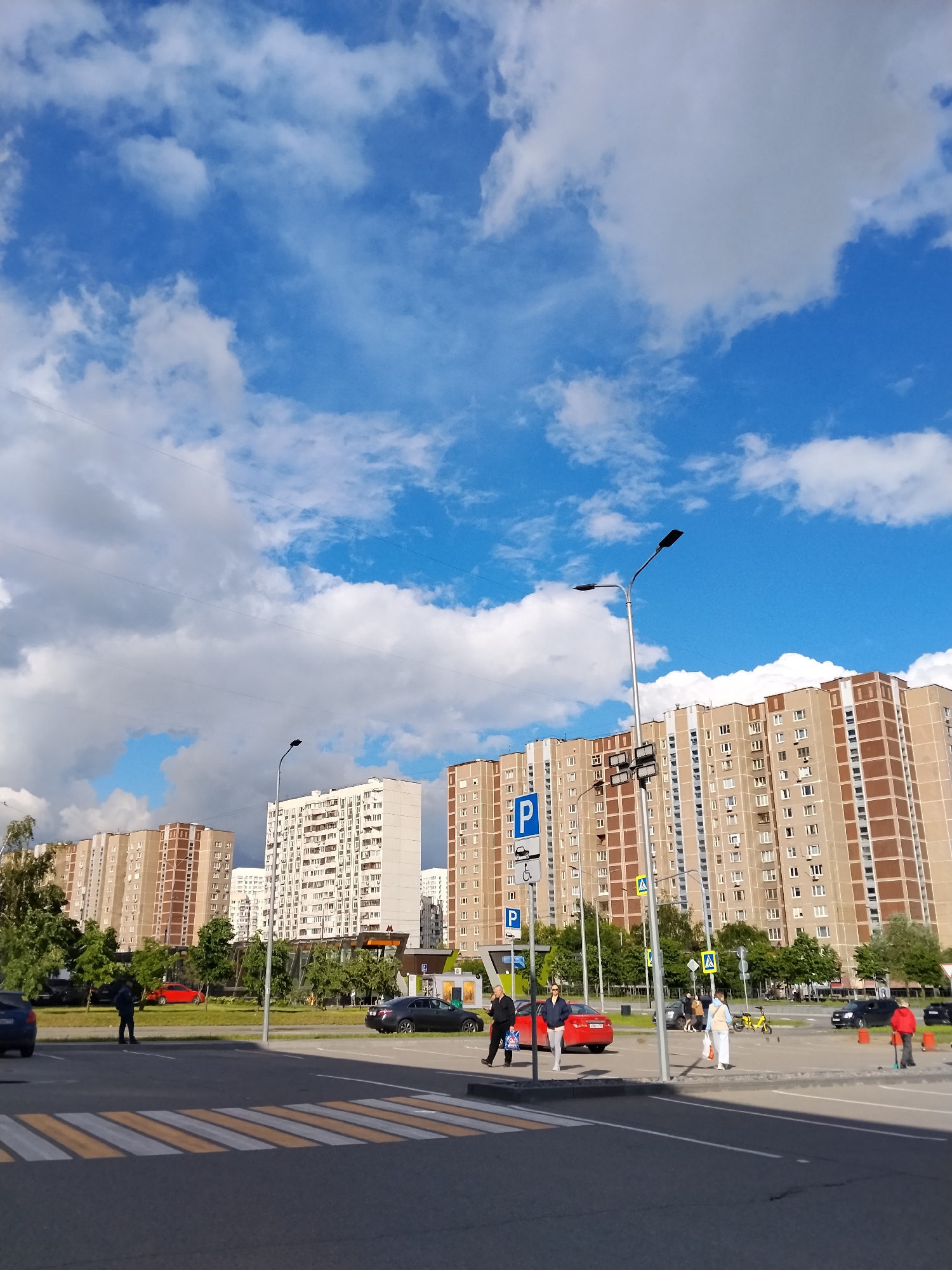The image depicts a typical urban scene in Moscow, Russia, under a partly cloudy sky. In the foreground is an asphalt road marked with a yellow and white pedestrian crossing. People are walking across the street and standing at the crosswalk. Cars of various colors, including black, red, and blue, are parked or driving nearby. In the background, several high-rise apartment buildings are visible, constructed in a style common to Moscow's residential areas. There is street lighting and parking signage. Trees are scattered around the parking areas and pedestrian zones.