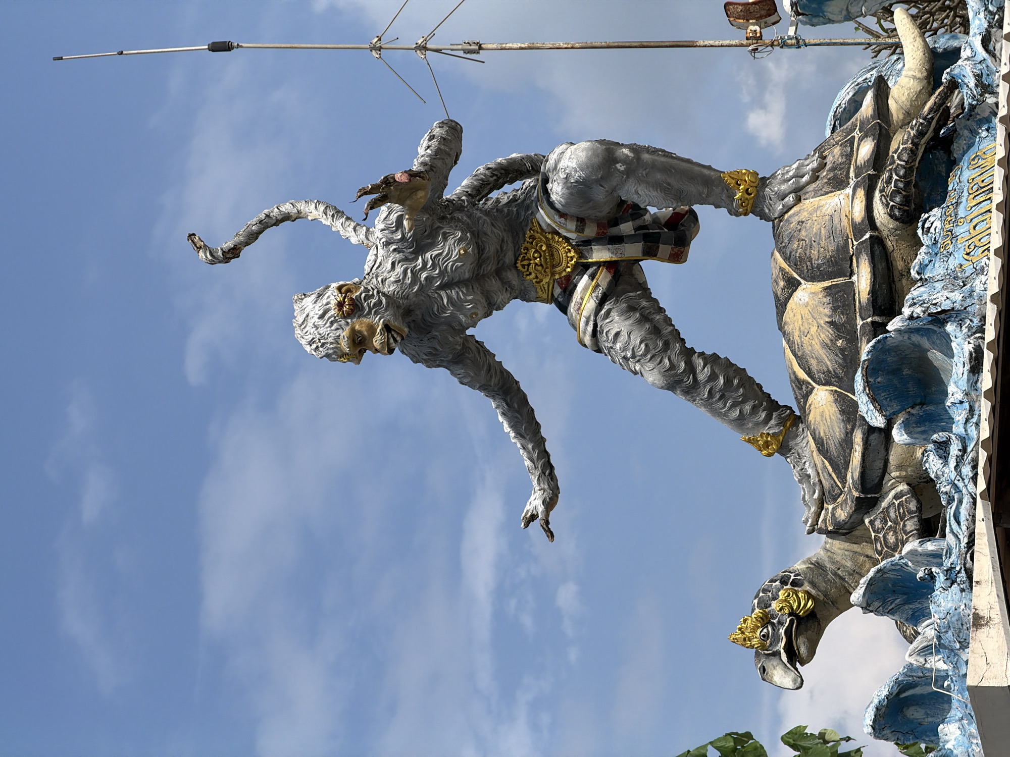 This is a medium shot, eye-level, outdoor photograph of a statue of a monkey-like creature standing on a turtle. The statue is grey with gold accents. The monkey has a distressed expression and its arms are outstretched, with one hand reaching forward. It is wearing a patterned sash around its waist. The turtle is also grey and black with gold decorative elements on its head and shell. The statue appears to be on a blue and white base that resembles waves. In the background, there is a bright blue sky with some scattered white clouds. A metal antenna is visible on the right side of the image. The statue is located in the city of Kuta Selatan, Indonesia.