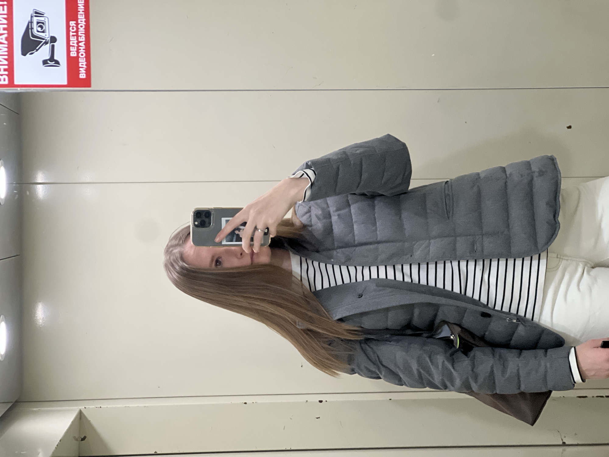 A woman is taking a selfie in an elevator. She is wearing a striped shirt, white pants, and a grey puffer jacket. Her hair is long and blonde. In the upper left corner of the image, there is a sign with a camera symbol and Russian text: "Внимание! Ведется видеонаблюдение" (Attention! Video surveillance is in progress). The elevator has off-white walls. The location is likely within a building in the city of Khimki, Russia, based on the language of the sign.