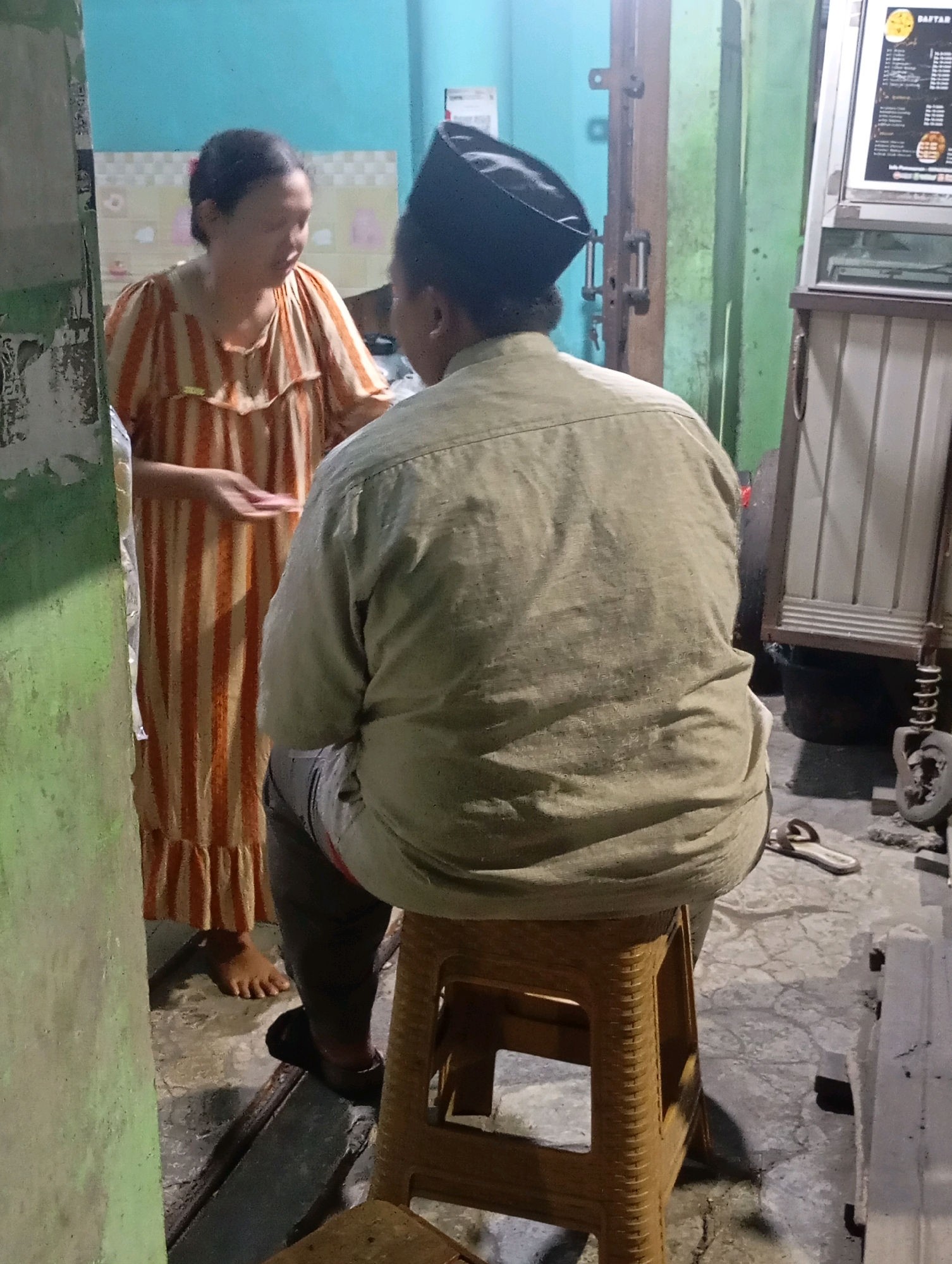 In Babelan, Indonesia, a woman in an orange and white striped dress stands talking to a man wearing a tan long-sleeved shirt and a black cap, who is seated on a tan plastic stool. The woman's hair is pulled back. Her right hand is extended. The man is facing away from the camera, his back to the viewer. Behind them, a door with metal hardware is visible, along with a green wall and a blue wall. To the right, a display board with text and images is mounted on a white and brown structure. The ground is concrete, with some debris and a pair of flip-flops visible.