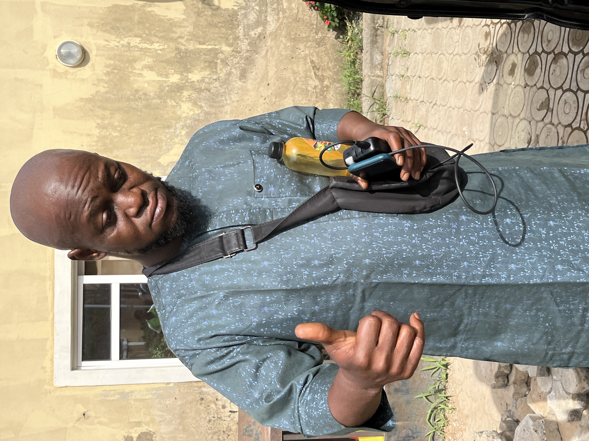 This image captures a man standing outdoors in Abuja, Nigeria, during the daytime. He is positioned against a light yellow or tan-colored building wall, which features a round, metallic light fixture and a white-framed window. The ground is paved with sections of decorative circular patterns and plain concrete, with small green plants growing along the edges.
The subject is a man with dark skin, a shaved head, and a beard. He wears a teal or dark green long-sleeved tunic, likely a traditional Nigerian attire, patterned with subtle light-colored floral or leaf motifs. A black shoulder or sling bag is worn across his body. His expression is neutral, looking towards the camera with a slight squint, indicative of bright sunlight.
In his left hand, the man holds a yellow plastic bottle, possibly containing a beverage, along with electronic devices that appear to be a mobile phone and a power bank or charger connected by cables. His right hand is raised slightly, with the palm open, suggesting a gesture in conversation or emphasis. The strong sunlight casts visible shadows, confirming it is a bright day. No discernible text is visible on any objects in the image.