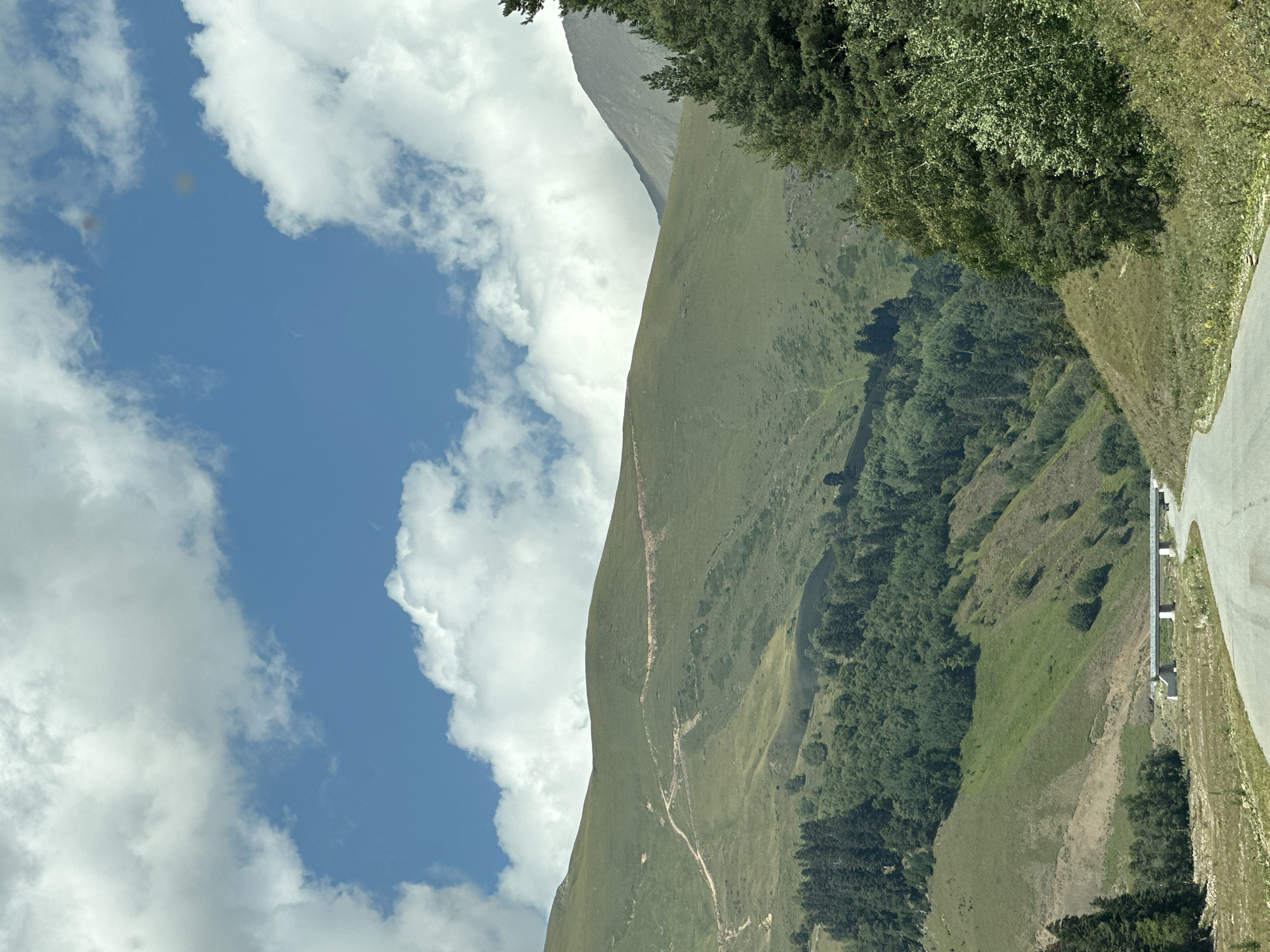 The image shows a scenic mountain landscape under a partly cloudy sky. In the foreground, a paved road curves to the right, leading into the distance. On the left side of the road, dry grass and sparse vegetation cover the ground. Further back, a wide, green mountain slope rises, with a winding dirt track visible on its surface. Below this slope, a dense forest of dark green trees covers a lower hill. A concrete bridge spans a gap in the middle ground, with a small, dark vehicle on it. The background features more rolling green hills and a taller, rocky mountain peak on the right, partially obscured by trees. The sky is blue with scattered white, fluffy clouds. This scene is located in Lunnaya Polyana, Russia.
