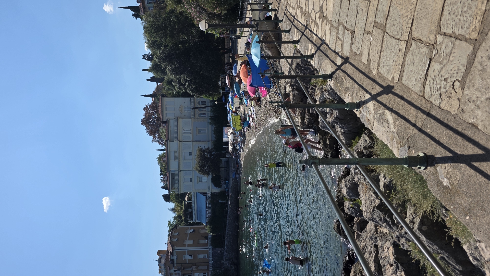 The image depicts a sunny day at a rocky beach on the Adriatic Sea. In the foreground, a stone walkway with a metal railing runs along the coast, casting long shadows. The sea is calm, with many people swimming and playing in the water. Some are floating on inflatable devices, while others are wading or swimming.
The beach itself is covered in pebbles and a few patches of sand. Numerous colorful umbrellas and beach chairs are set up, indicating a popular spot for sunbathing and relaxation. A few individuals are lying on towels, while others are seated under the shade of umbrellas.
In the background, a row of buildings lines the coast. These structures appear to be residences or perhaps small hotels, with some featuring balconies and tiled roofs. Lush green trees and vegetation are interspersed among the buildings, adding to the scenic coastal atmosphere. The sky is a clear, bright blue with a single, small white cloud. The overall scene conveys a sense of leisure and enjoyment on a warm summer day.