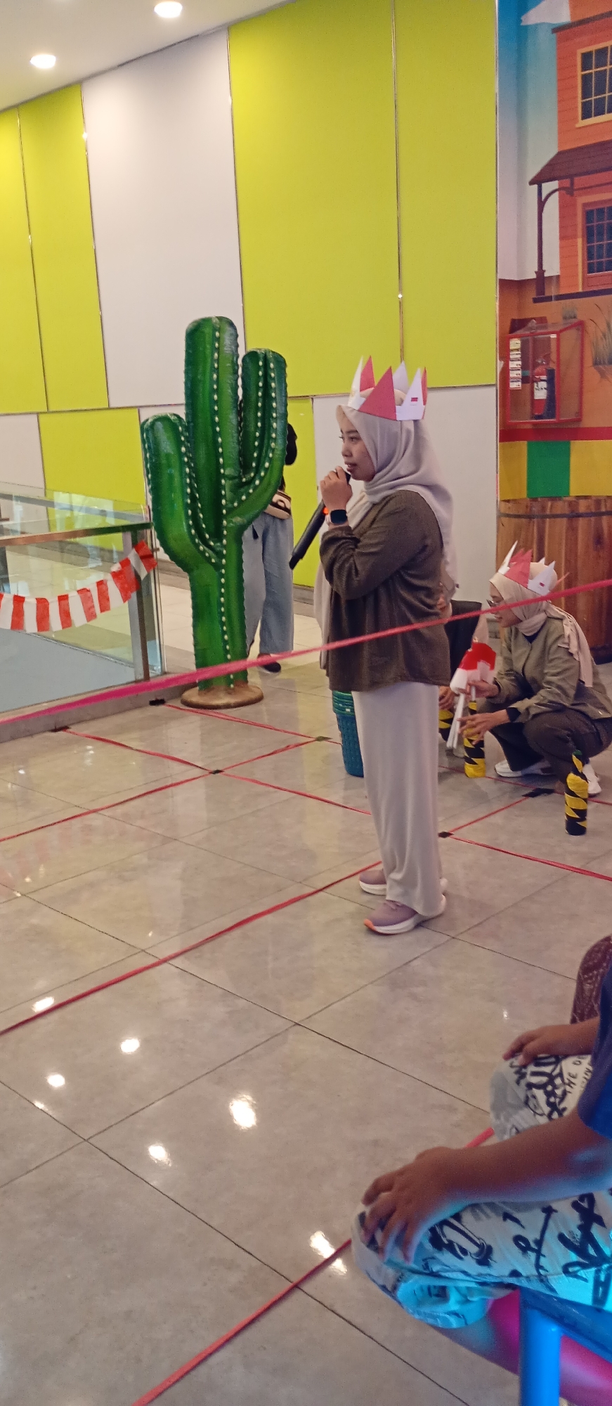 The image depicts an indoor event, likely a themed celebration or competition, with a vibrant backdrop. In the foreground, a large, green, artificial cactus stands prominently. A woman wearing a light brown hijab and a dark green long-sleeved shirt is holding a microphone and speaking, wearing a paper crown. To her right, another person, also wearing a paper crown and a similar outfit, is kneeling. The floor is tiled with a grid pattern, and red ribbons are laid out on the floor in a square formation, suggesting a game or activity area. In the background, a mural of a Wild West-themed building is visible, along with a fire extinguisher. The walls are painted in alternating sections of bright yellow and white. The overall impression is of a festive and engaging event. The location is Probolinggo, Indonesia.