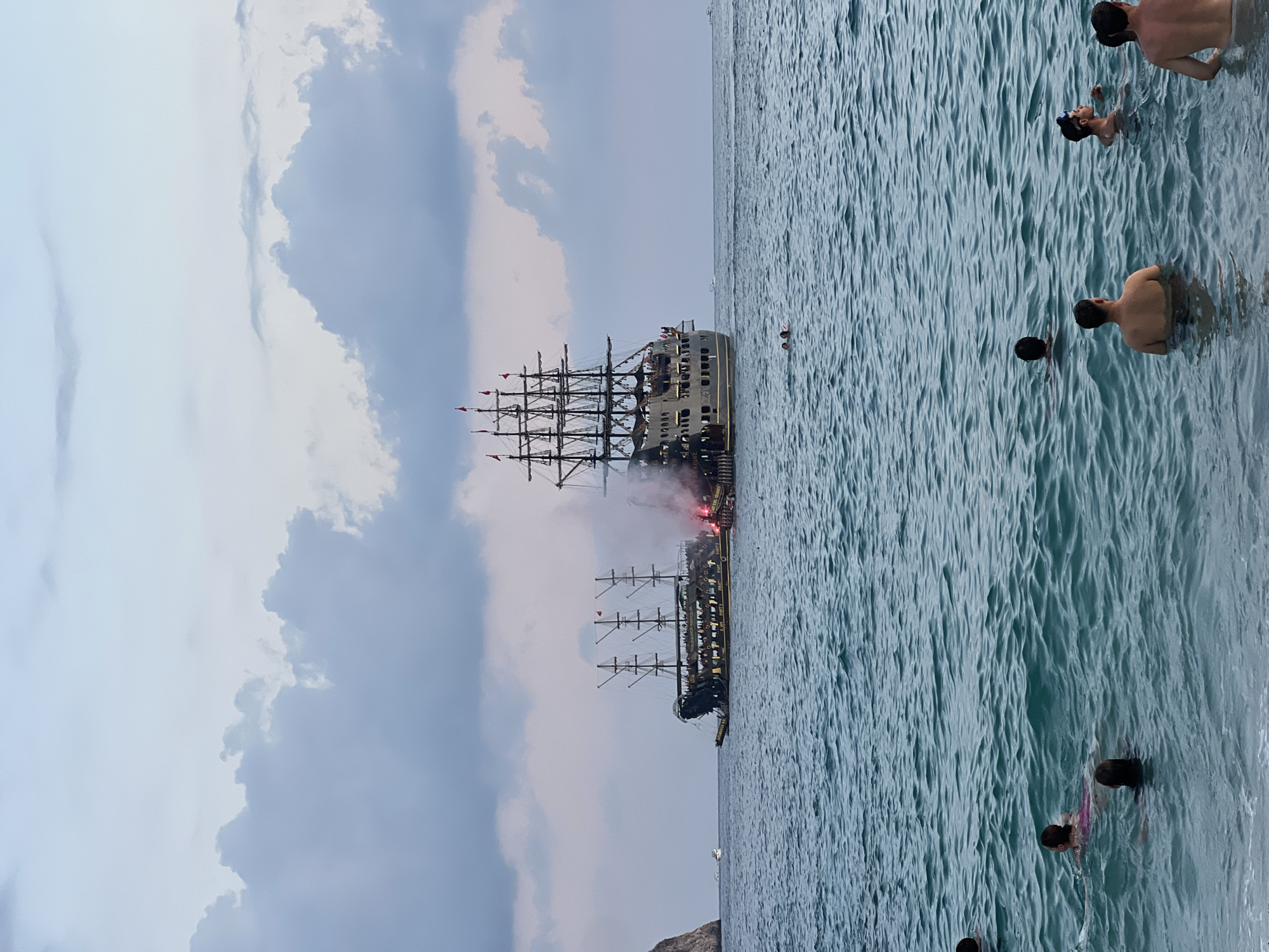 The image shows a large, elaborately decorated pirate ship sailing on the sea with people on its deck and in the water. The ship is adorned with multiple masts and sails, and it appears to be part of a "Pirate Party" as indicated by text on the ship's hull. Red flares are visible, emitting smoke, suggesting a celebratory event or a theatrical performance. In the foreground, several people are swimming and wading in the clear blue water. In the background, a rocky cliff face is visible on the left side of the frame, characteristic of the coastline of Alanya, Turkey. The sky is overcast with soft clouds, indicating either dawn or dusk. The overall scene suggests a tourist attraction or a special event taking place in the waters off Alanya.