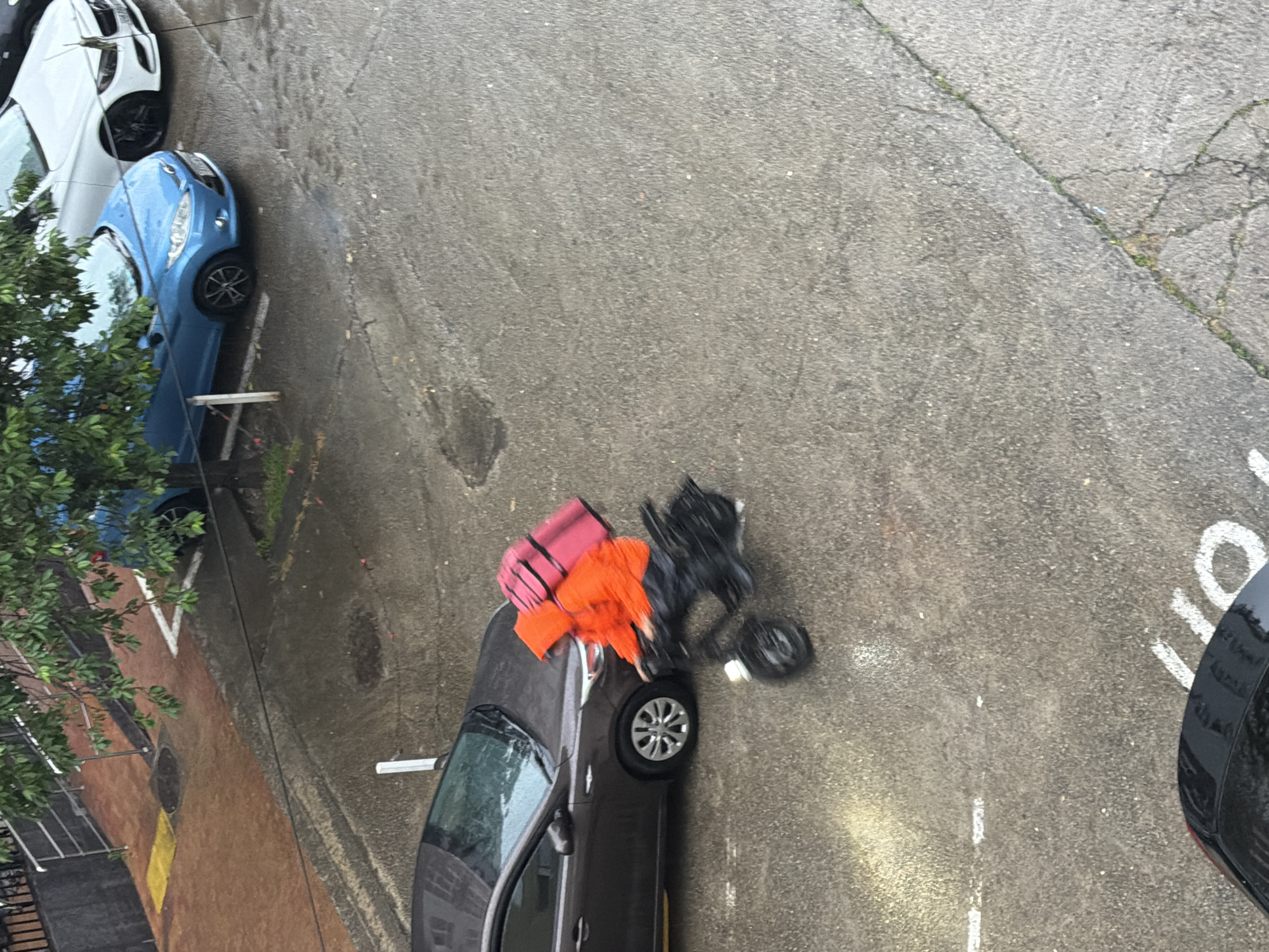 An aerial view captures a blurred motion of a person on a scooter in a wet, urban environment. The individual is wearing an orange hooded jacket and black pants, with a large pink bag strapped to their back. They are riding past a dark-colored car parked on a wet asphalt surface. In the background, a blue car and a white car are visible, also parked. The scene appears to be a street or parking lot, with a sidewalk area featuring red paving stones and a tree with green foliage. The overall impression is of a rainy day in a city, with the motion blur indicating the scooter's movement. This scene is consistent with typical urban street views found in Sochi, Russia, particularly during inclement weather.