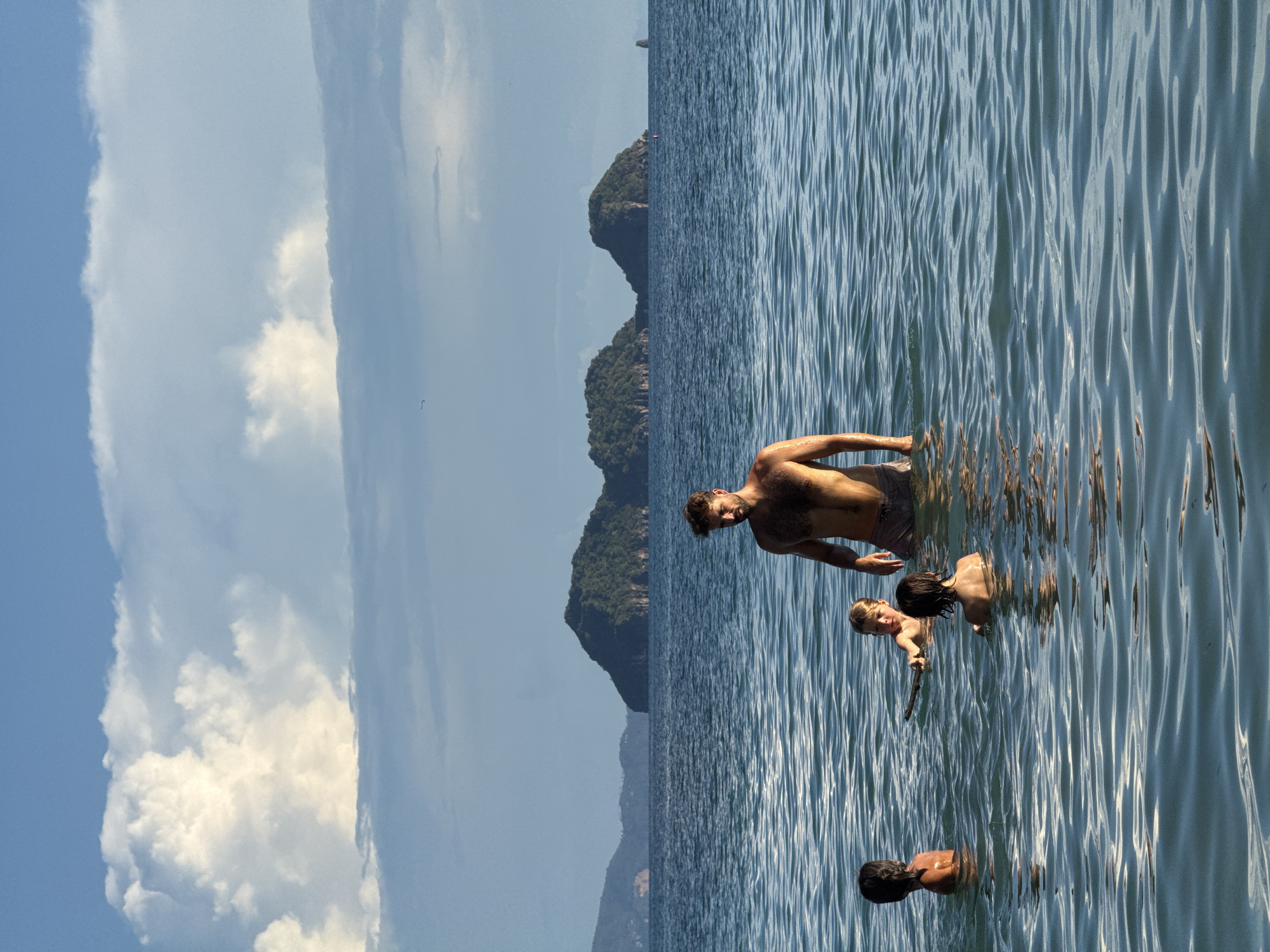 A group of people, including a shirtless man with dark hair and a beard and at least three children, are wading in the ocean. The man stands in the foreground, facing left, while the children are positioned to his left and in front of him. One child, with light hair, holds a stick. The water is calm with ripples, reflecting the sky. In the background, several green, tree-covered islands are visible in the distance, rising from the sea under a partly cloudy blue sky. The scene is set in ตำบลเกาะพะงัน, Thailand, likely during daylight hours with the sun casting a warm glow on the water and figures.