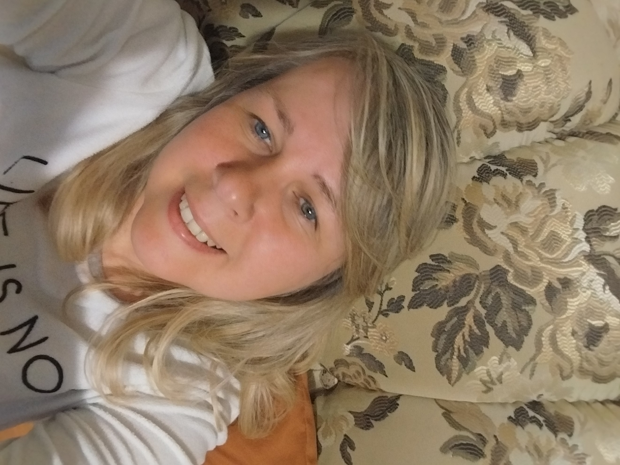 A close-up, tilted selfie captures a blonde woman with blue eyes, smiling warmly at the camera. She appears to be lying down, her head resting on a patterned cushion or sofa upholstery featuring large floral motifs in shades of beige, brown, and off-white. Her long, light blonde hair frames her face and spills onto the background. She is wearing a white long-sleeved top, on which partial black text is visible, reading "IS NO O". Her bright smile reveals white teeth, and her expression conveys happiness and relaxation. A glimpse of a solid orange or brown cushion is visible near her lower left side. The lighting suggests an indoor setting, possibly daytime or a brightly lit room. The location context provided is Orenburg, Russia.