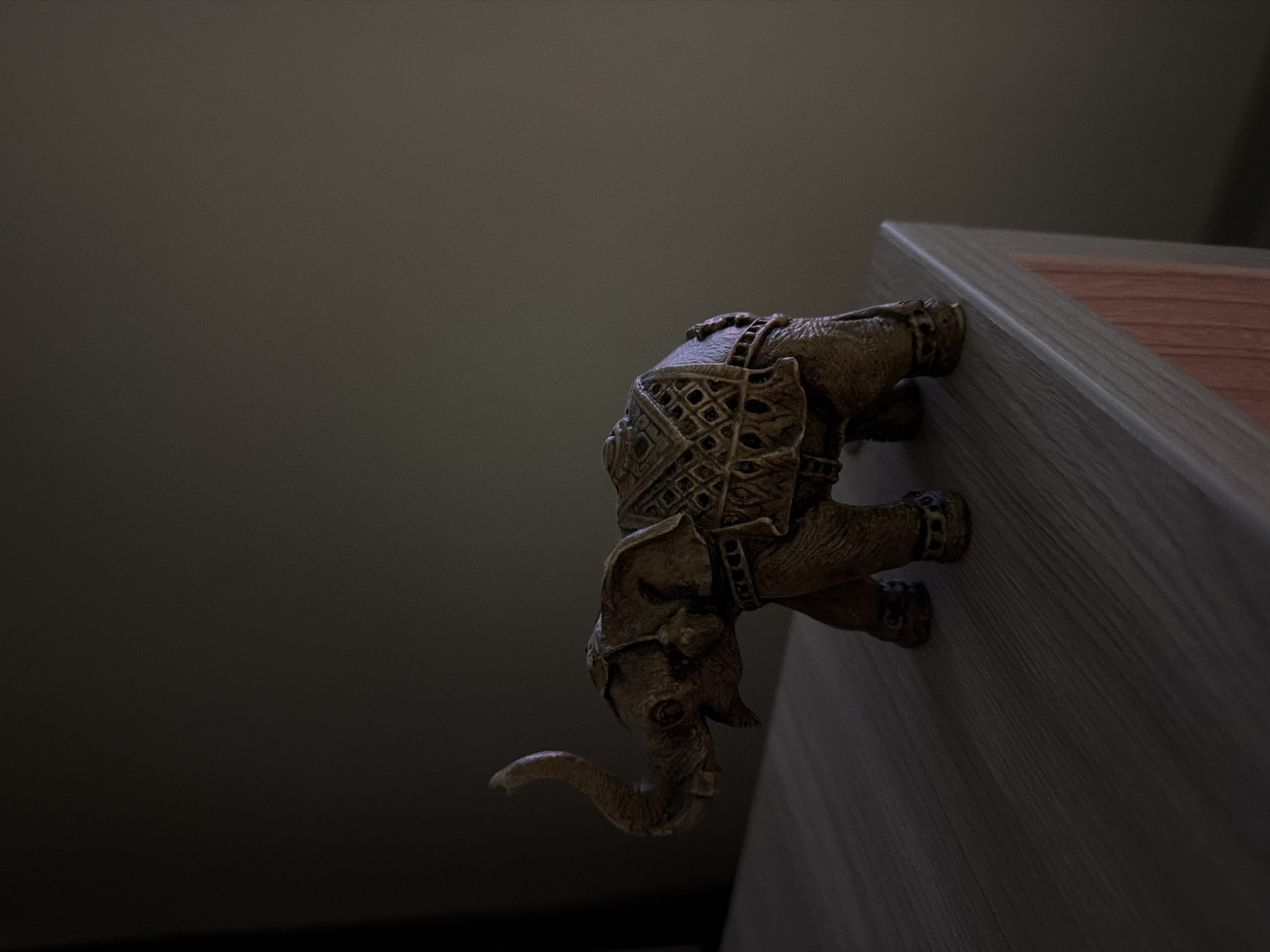 A decorative elephant figurine sits on a wooden surface, possibly a shelf or table, in a dimly lit room. The figurine is made of a bronze-colored material and is detailed with ornate saddle-like adornments. Its trunk is raised in the air. The background is a plain, dark wall, and the lighting creates a moody atmosphere with shadows playing across the scene. The surface the elephant is on has a wood grain texture. The location in the city of Naberezhnye Chelny, Russia, is not discernible from the image.