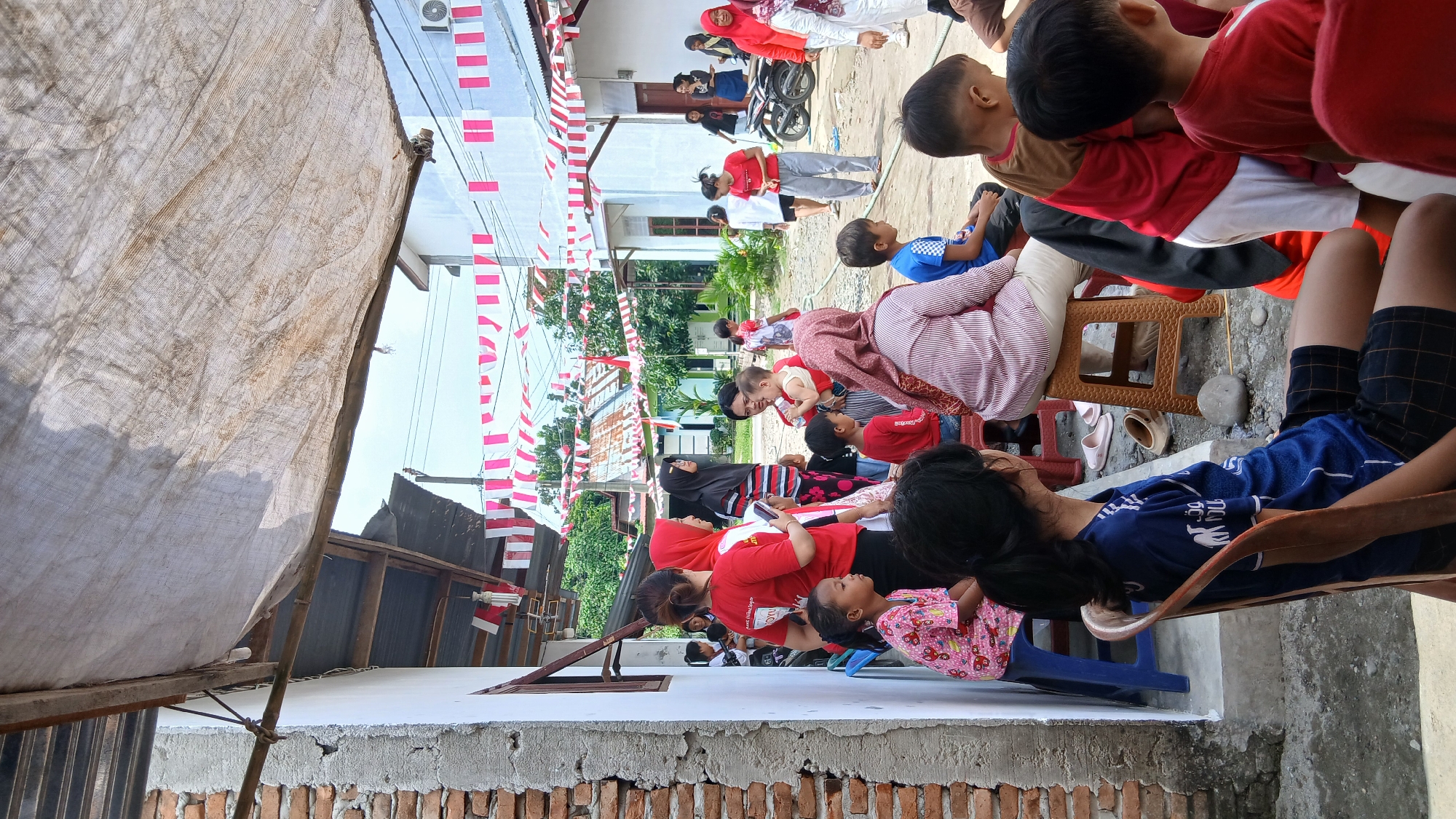 In Klambir Lima Kebun, Indonesia, a gathering of people is taking place outdoors. The scene is decorated with red and white Indonesian flags strung across the area, suggesting a national celebration or event. A weathered, light-colored tarp is stretched overhead, providing partial shade. In the foreground, a young person in a blue t-shirt and plaid shorts is seated on a wooden chair, looking to the right. Behind them, a person wearing a pink and white striped shirt is seated on a brown stool. To their left, a young girl in a pink patterned dress sits on a blue chair. Several other individuals, including adults and children, are visible in the background, some interacting and others observing. A motorcycle is parked in the middle ground. The environment appears to be a residential or community area with buildings and greenery visible.