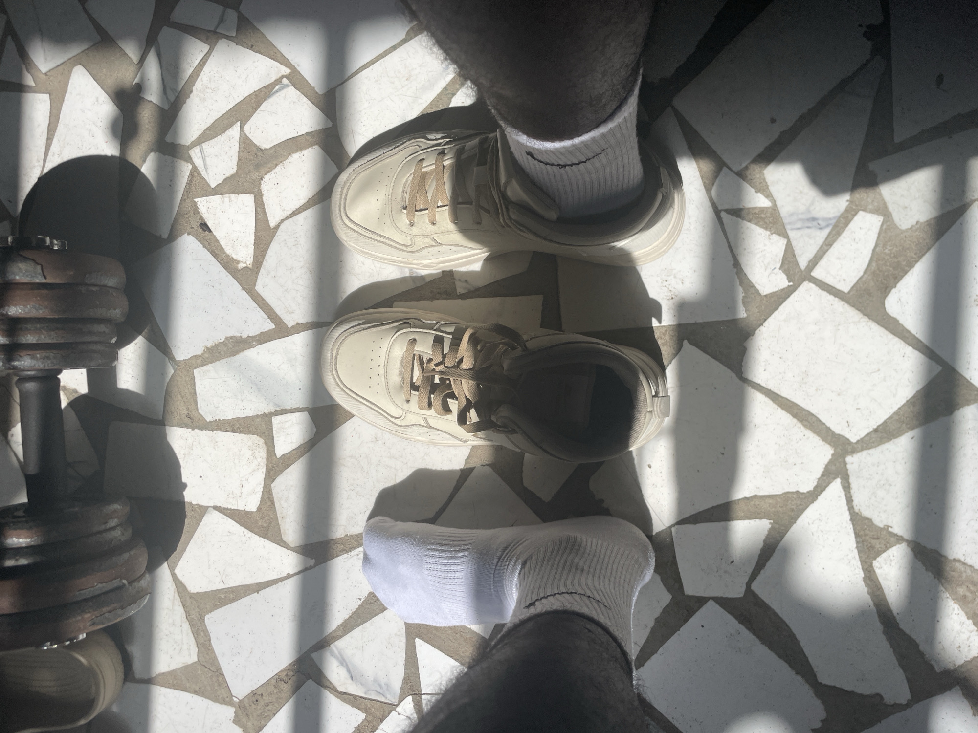 The image presents a top-down view of a person's lower legs and feet, set against a backdrop of a distinctively tiled floor. The person has dark-skinned, hairy legs and is wearing white athletic socks. The left foot is inside a light beige or off-white sneaker with matching laces, while the right foot is bare, clad only in a white sock, with its corresponding sneaker placed beside it on the floor. A visible Nike swoosh logo is present on the white sock of the right foot.
To the left of the person's feet, a dark, rusty dumbbell with several weight plates is resting on the floor. The flooring consists of irregularly shaped white or light-colored tiles embedded in dark gray grout, creating a mosaic or terrazzo-like pattern.
The scene is illuminated by bright, direct sunlight, which casts long, sharp linear shadows across the floor, suggesting light passing through a grille, window bars, or a similar structure. These strong shadows indicate a clear, sunny day, likely during midday or early afternoon. The overall setting appears to be an outdoor or semi-outdoor area, potentially a home workout space in Badagry, Nigeria, as suggested by the location context. The presence of athletic footwear and a dumbbell implies an activity related to exercise or physical training. No emotions are discernible.