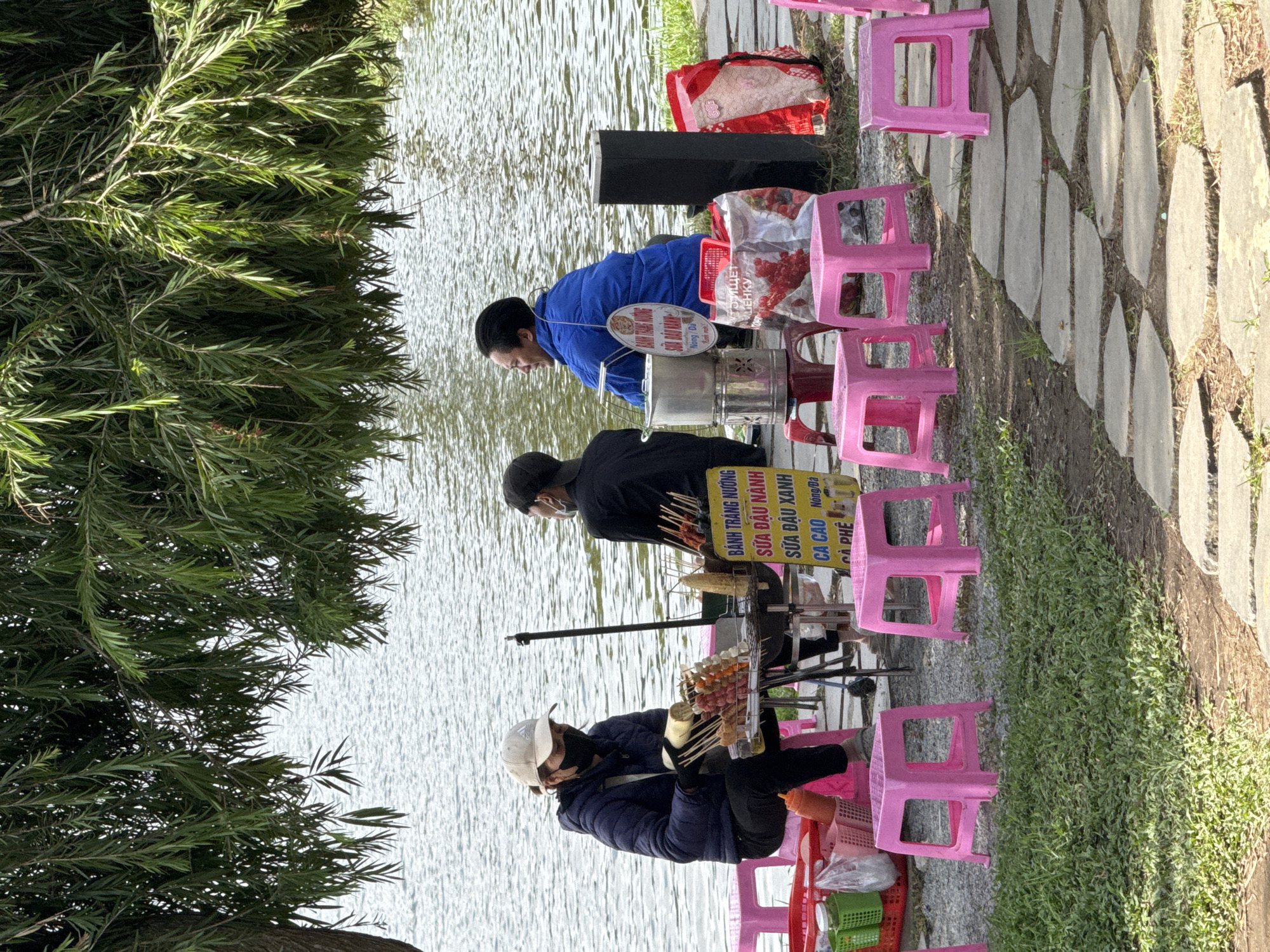 The image depicts a street food vendor scene by a lake, likely in Da Lat, Vietnam, given the foliage and general ambiance. Two individuals, a woman and a man, are present. The woman, wearing a dark blue jacket, black pants, and a white baseball cap, is seated on a pink plastic stool. She is wearing a black face mask and gloves and is in the process of preparing food, holding a skewer with what appears to be meat and sauce. In front of her is a grill with skewers of food and a sign with Vietnamese text.
To her right, a man in a black jacket and dark pants is seated on another pink plastic stool, facing away from the camera and looking towards the lake. He has a black mask on his face. Further to the right, a woman in a bright blue jacket is seated, also facing away from the camera and looking at the lake. She is wearing a white sign around her neck.
The foreground is characterized by a path made of large, irregularly shaped stone slabs, with patches of grass and more pink plastic stools. In the background, a body of water, presumably a lake, stretches across the scene, with the gentle ripples suggesting a calm environment. Overhanging branches of a large tree, with long, slender green leaves, provide shade for the vendor and her customers. Several bags and containers are visible near the vendor's stall, including a red plastic basket and a large red and white patterned bag. A dark rectangular object, possibly a trash bin or speaker, stands to the right of the seated man. The overall scene conveys a relaxed, outdoor atmosphere typical of a lakeside setting where vendors offer local delicacies.