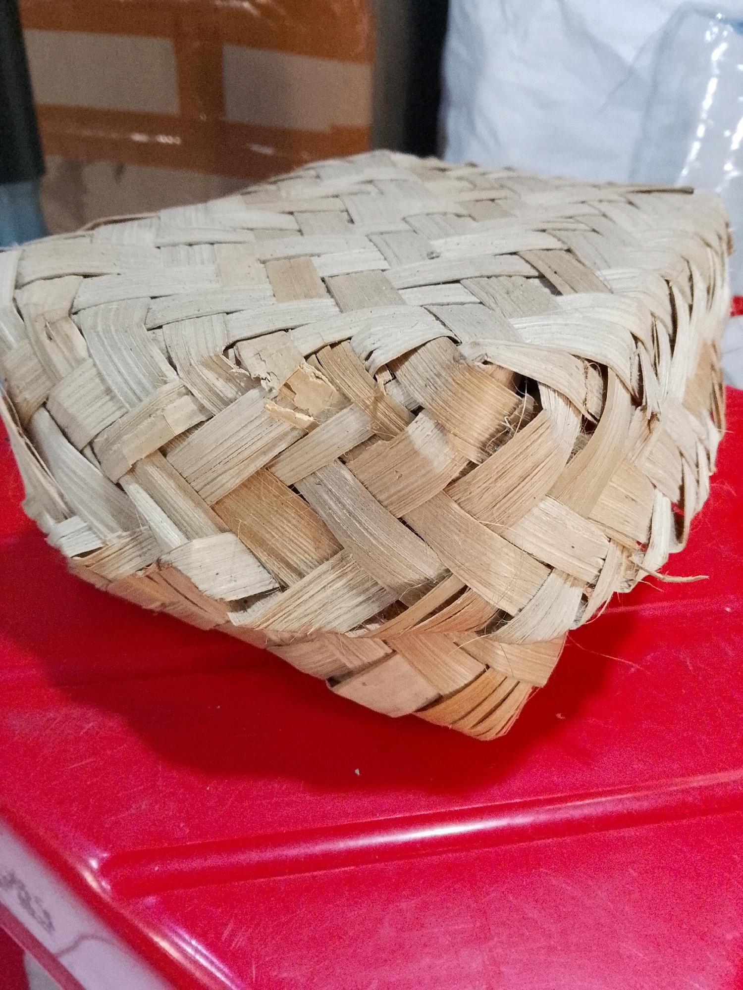 A woven basket made of bamboo or similar material sits on a red surface. The basket has a square base and its sides are woven in a diamond pattern. The material appears dry and aged, with some strands frayed. In the background, out-of-focus objects include cardboard boxes and white packaging. The red surface is textured with some visible scratches and dust. The lighting suggests an indoor environment, possibly a storage area or a shop in Surabaya, Indonesia. There are no people or animals visible in the image.