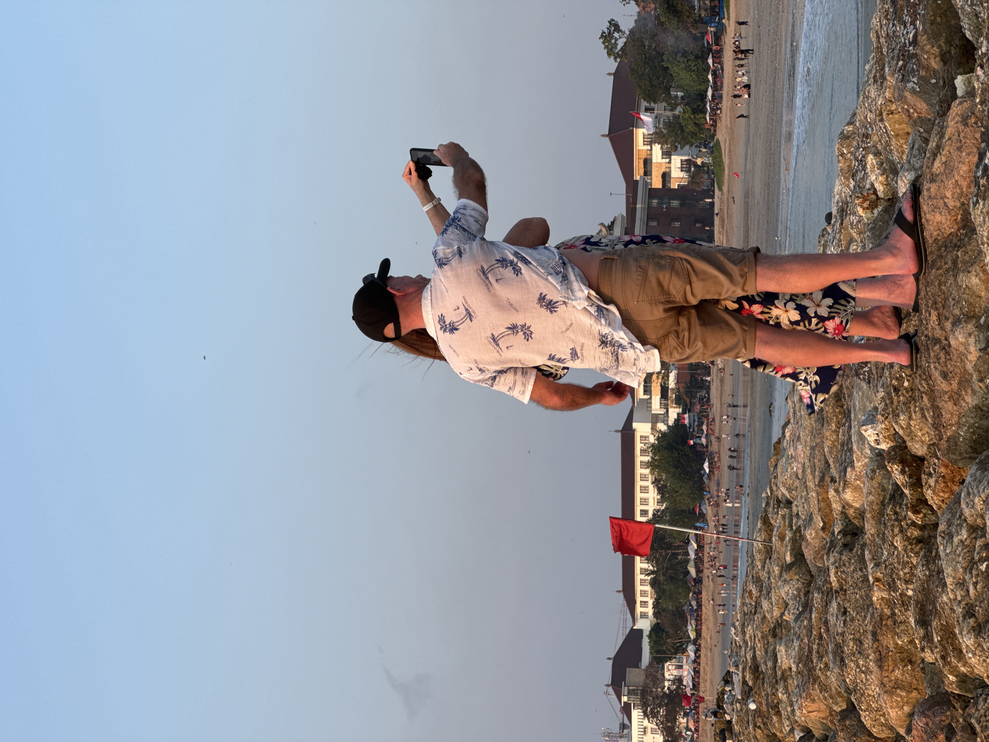 The image captures a couple standing on a rocky breakwater overlooking Kuta Beach in Bali, Indonesia. The man, wearing a Hawaiian-style shirt and khaki shorts, is holding a smartphone to take a photograph. A woman, whose legs and floral skirt are visible, stands beside him. The background reveals a bustling beach scene with numerous umbrellas and people enjoying the sun and sea. A prominent red flag, signaling the sea conditions, stands on the breakwater, indicating potentially dangerous waters. Buildings line the coast, and the clear sky suggests it is late afternoon or early evening, with the setting sun casting a warm glow on the rocks. The overall atmosphere appears to be one of leisure and tourism.