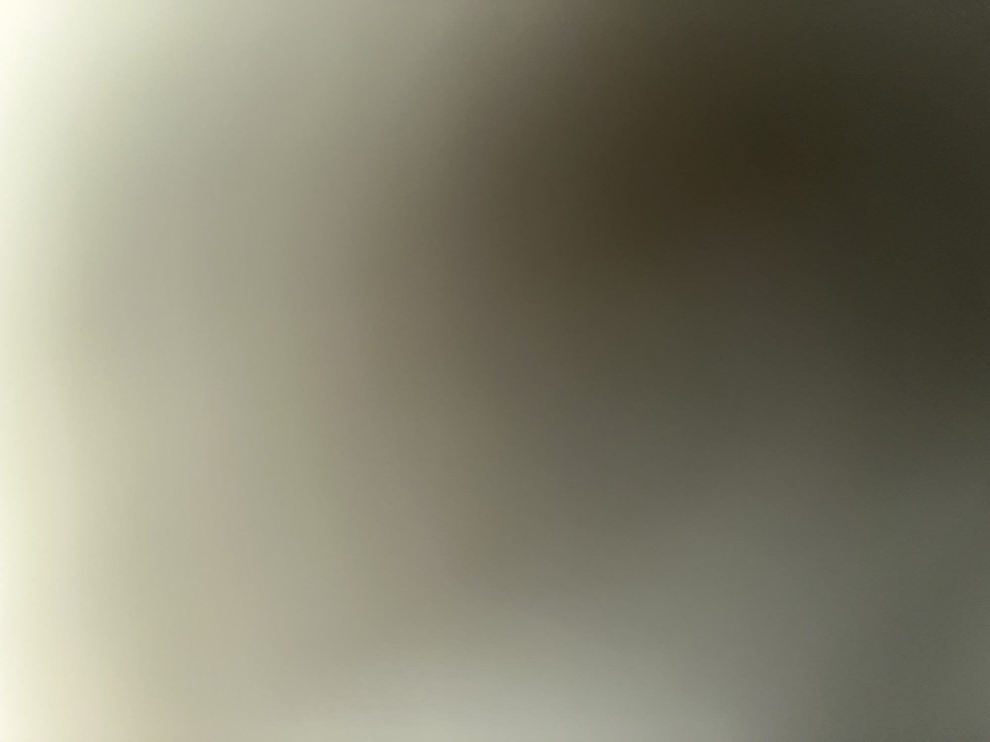 The image is a blurred, abstract representation of a wall or surface. There are no discernible people, objects, actions, or interactions. The lighting is soft and diffuse, creating a gradient of light and shadow across the surface. The colors are muted, ranging from a pale off-white at the top to a darker, desaturated brown at the bottom. There is no information in the image that can be used to determine its location in the city of Dubeczne, Ukraine, or any other specific location.