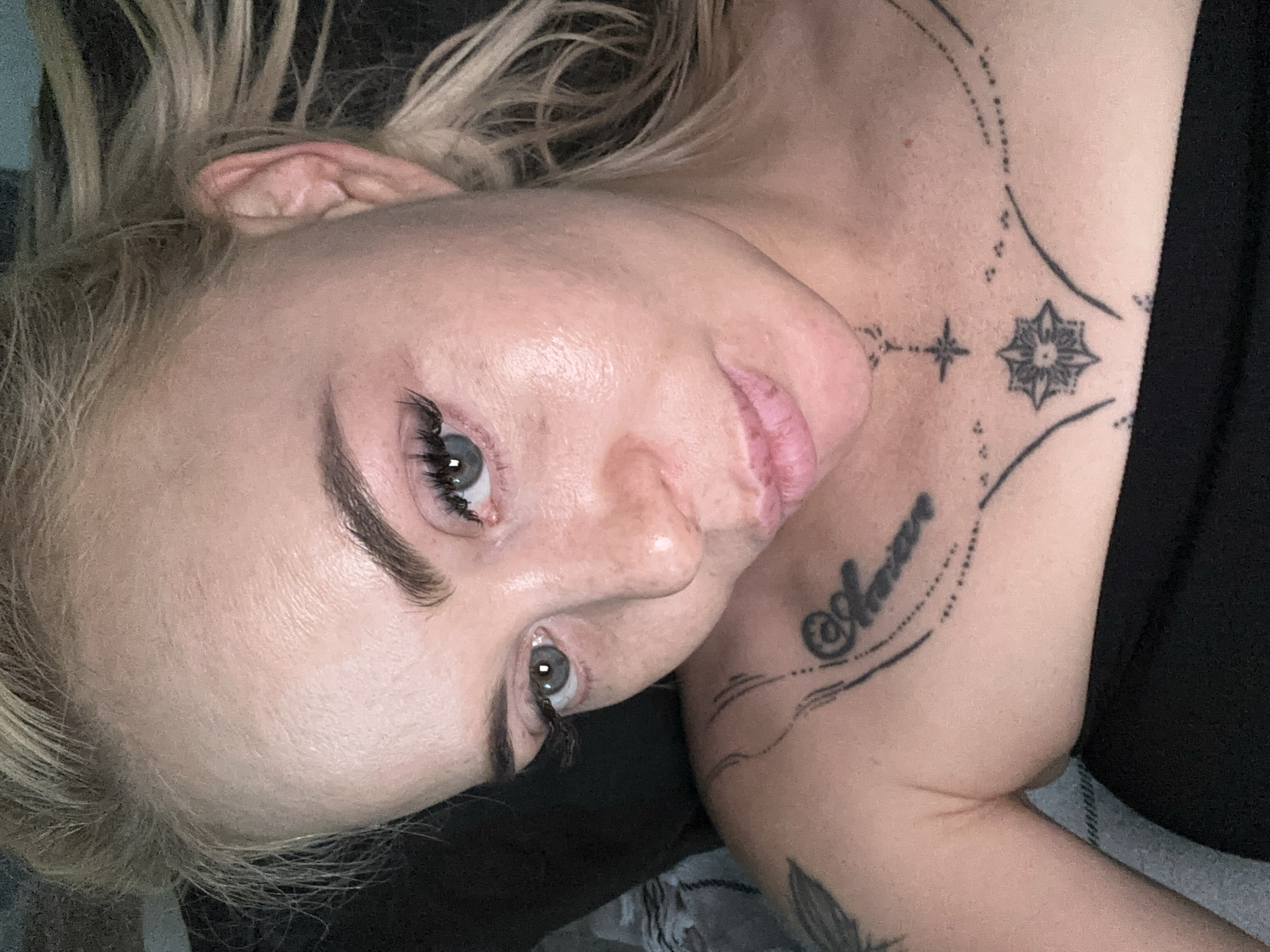 The image displays a close-up, slightly angled portrait of an adult female with fair skin, positioned centrally. Her blonde hair is styled back, with loose strands visible around her face and behind her right ear. She has light-colored eyes, prominent dark eyebrows, and long dark eyelashes. Her lips are full and slightly parted, showing a slight pout. Skin texture, including minor lines on her forehead and small bumps around her mouth, is visible. She wears a black strapless top or bandeau, visible across her chest and shoulders. Several black tattoos are present: a partial design on her left upper arm/shoulder, script lettering followed by a dotted line pattern on her left upper chest, and a prominent geometric star-like motif on her sternum, accompanied by smaller star/cross symbols and curving dotted lines that extend horizontally. The subject is looking directly into the camera. The background is dark and out of focus, with a faint grey textured surface partially visible on the left side, consistent with an indoor setting.
