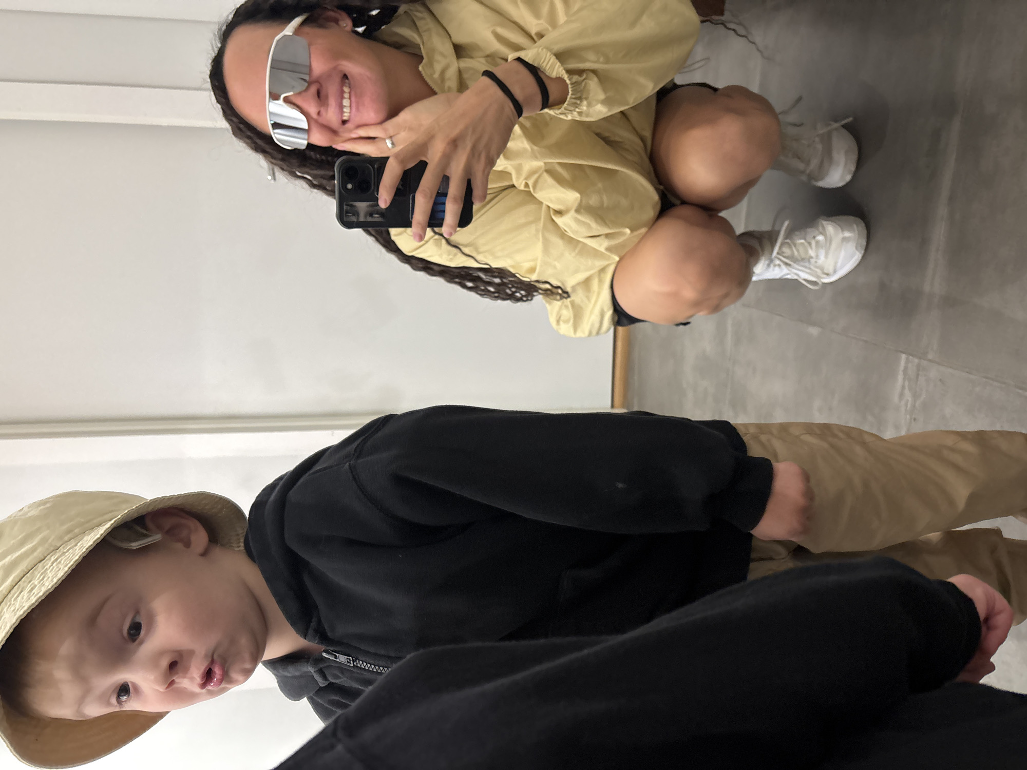 In an indoor environment with a light-colored wall and a grey tiled floor, a young child wearing a light-colored bucket hat and a black zip-up hoodie is positioned on the left side of the frame. The child has their mouth puckered and their eyes are looking to the left. On the right side of the frame, an adult with dark, braided hair is crouching and holding a black smartphone to take a mirror selfie. The adult is wearing oversized white sunglasses and a light yellow hooded jacket, with white athletic shoes. The adult is smiling and touching their chin with their left hand. The overall scene appears to be a casual moment captured in a mirror. There is no information in the image to suggest the location is in Chaikovsky, Russia.