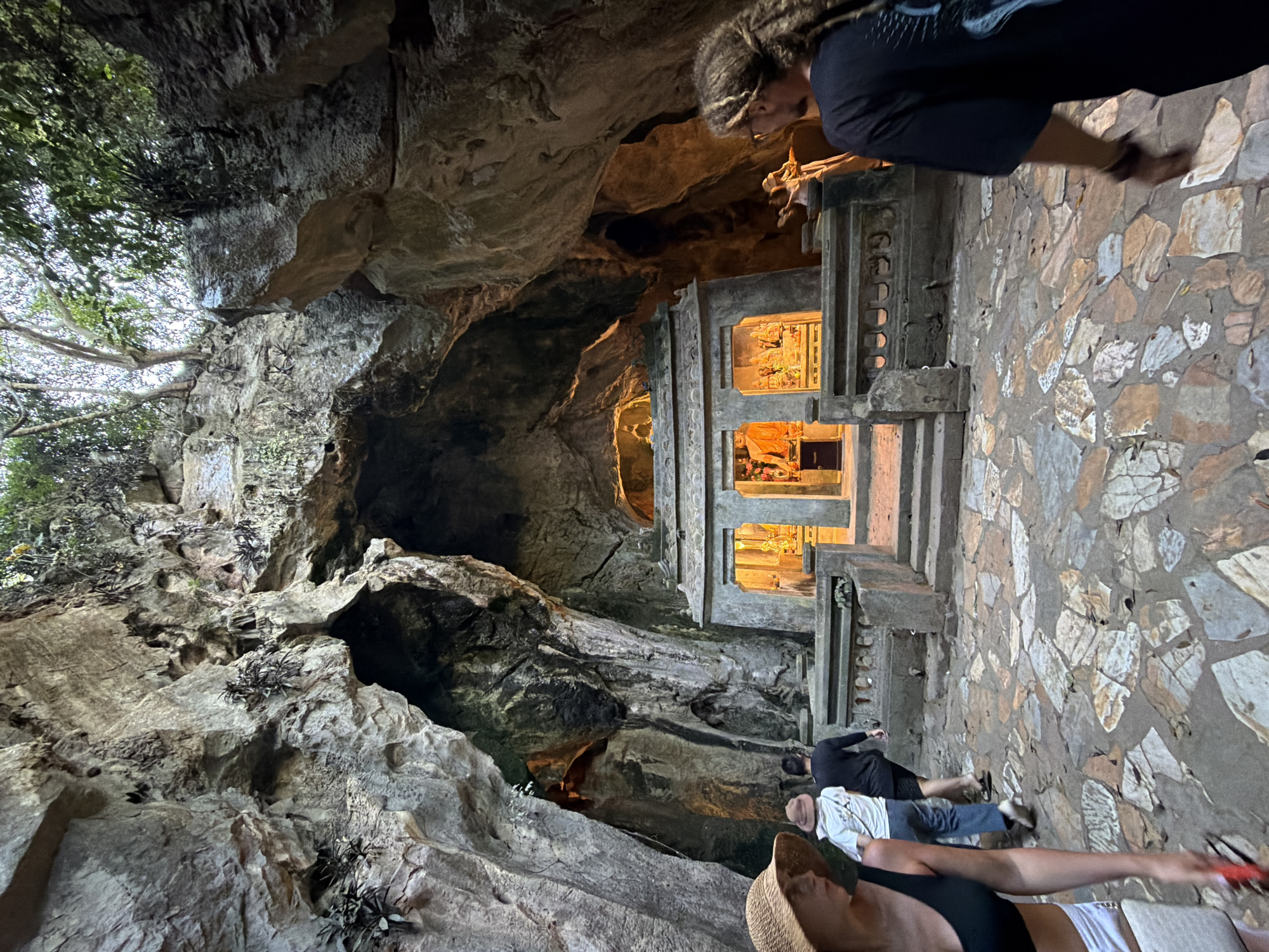 The image depicts the entrance of a cave, likely within the Marble Mountains in Đà Nẵng, Vietnam, featuring a Buddhist shrine. In the foreground, a woman wearing a straw hat and a black bikini top is partially visible on the left, facing towards the cave. To her right, a man wearing a beige hat and a white t-shirt walks away from the camera, followed by another man in a black shirt. The path leading to the shrine is paved with irregular, multi-colored stones. The shrine itself is an ornate structure with multiple levels, adorned with statues and lit from within. The cave's natural rock formations create a dramatic backdrop, with lush greenery visible at the opening. On the right side of the frame, a person with long, dreadlocked hair and a black t-shirt is visible, looking towards the shrine. The lighting suggests it is late afternoon or early evening, with the shrine illuminated by artificial lights.